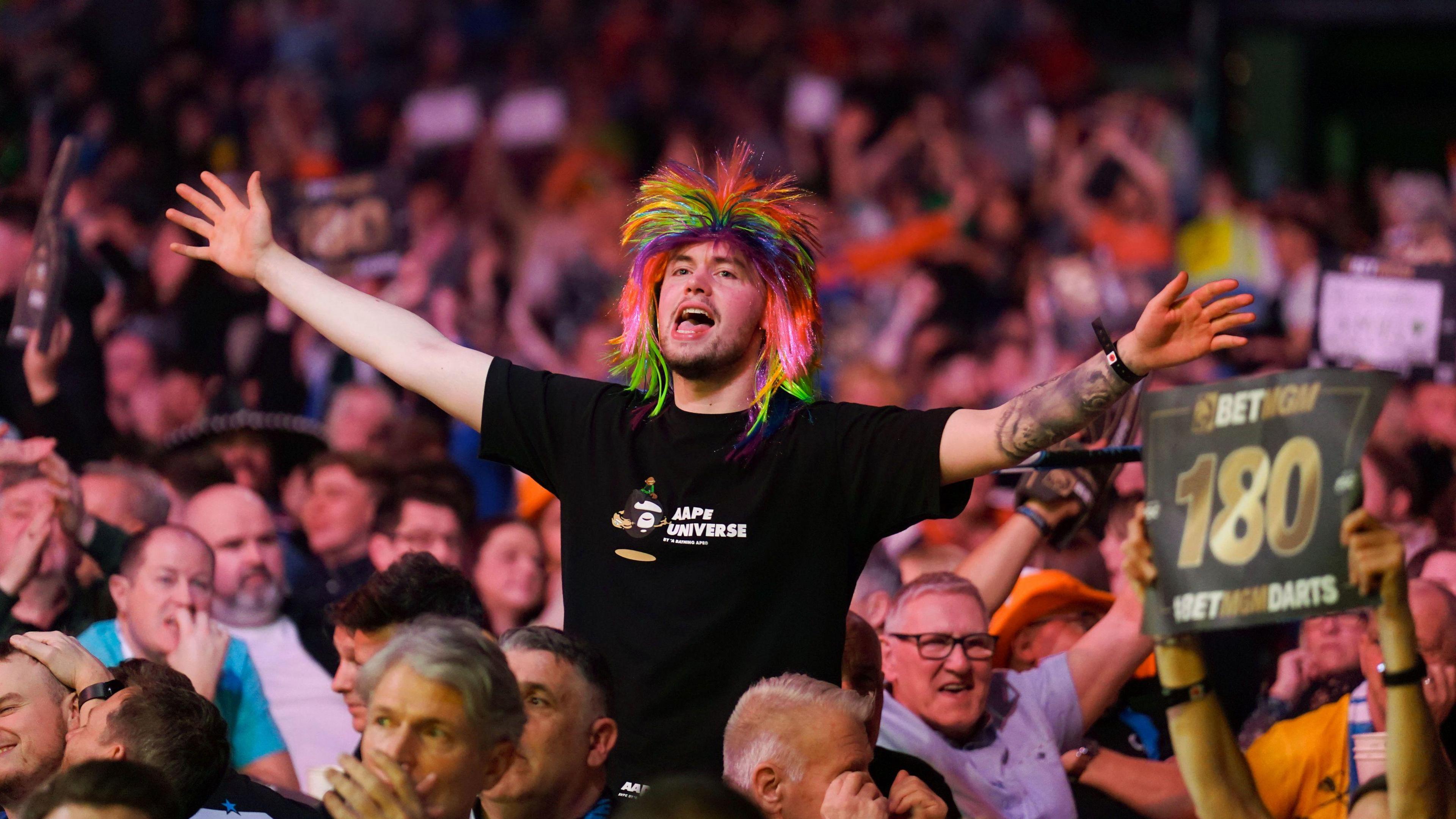 A crowd of people watching a darts contest at Glasgow's Hydro. A man in the middle of the crowd is on his feet, wearing a multi-coloured wig and a black t-shirt. His arms are out-stretched and his mouth open wide. He's enjoying himself.