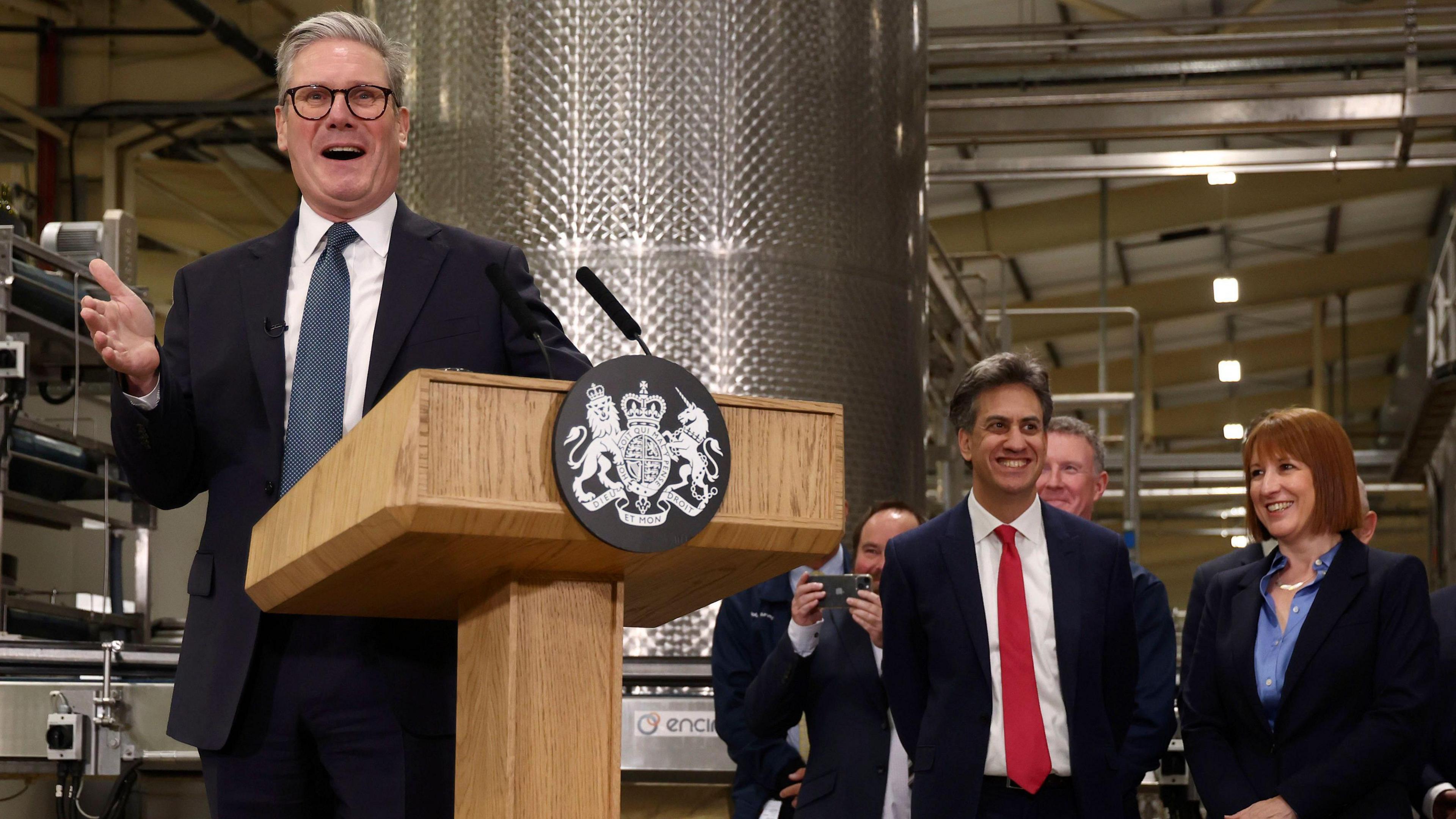 Sir Keir Starmer makes a speech in front of a wooden podium. He has his hand clenched. He is wearing a suit and navy tie. Rachel Reeves, in a blue shirt and suit jacket, and Ed Miliband, in a red tie and white shirt and navy suit, look on during a tour of a factory in Cheshire.