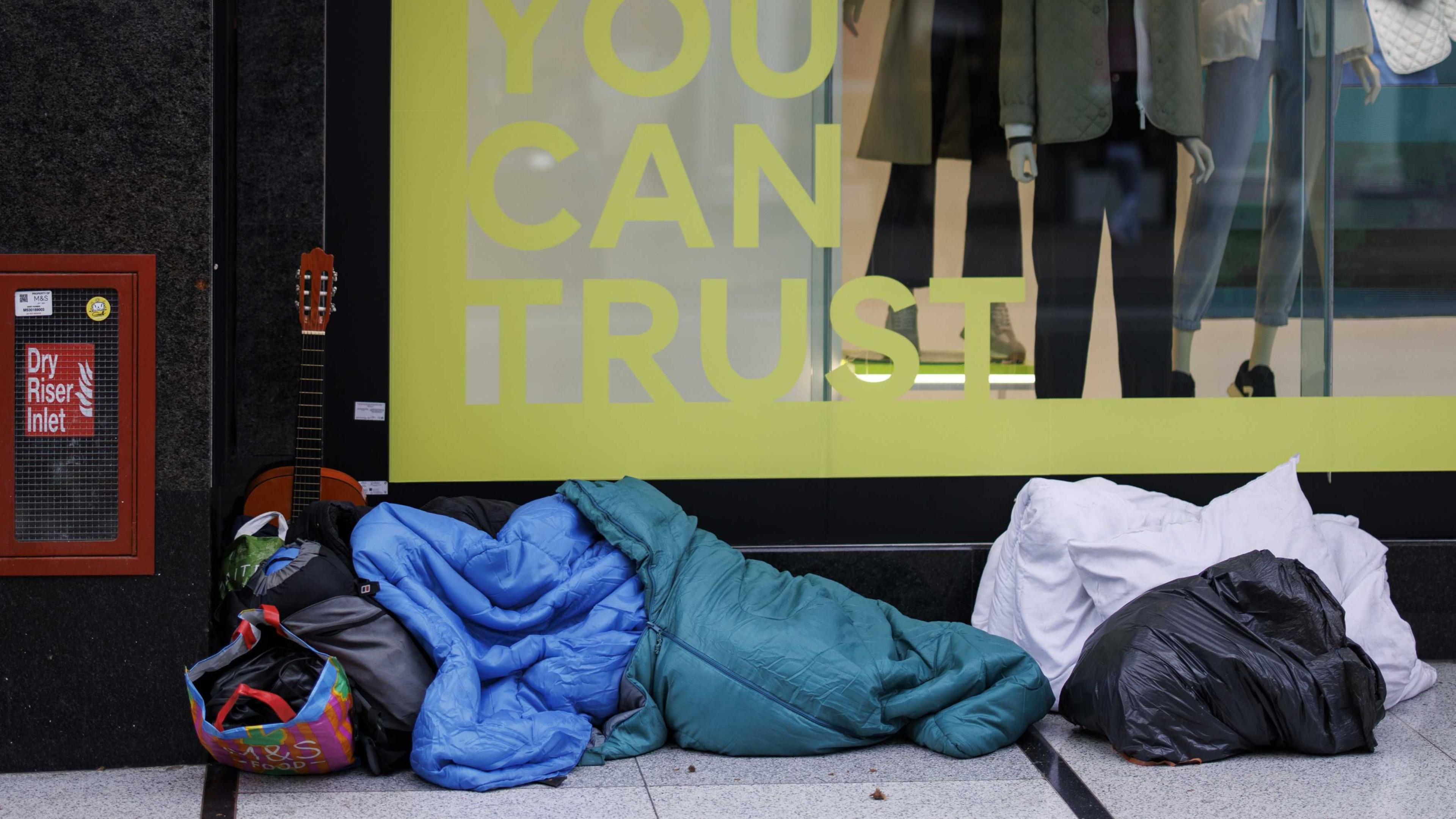 A person sleeping rough, entirely covered in sleeping bags, rests in front of a shop window on Oxford St. There are bags of different colours and sizes at their head and feet and a guitar propped against the window behind them.