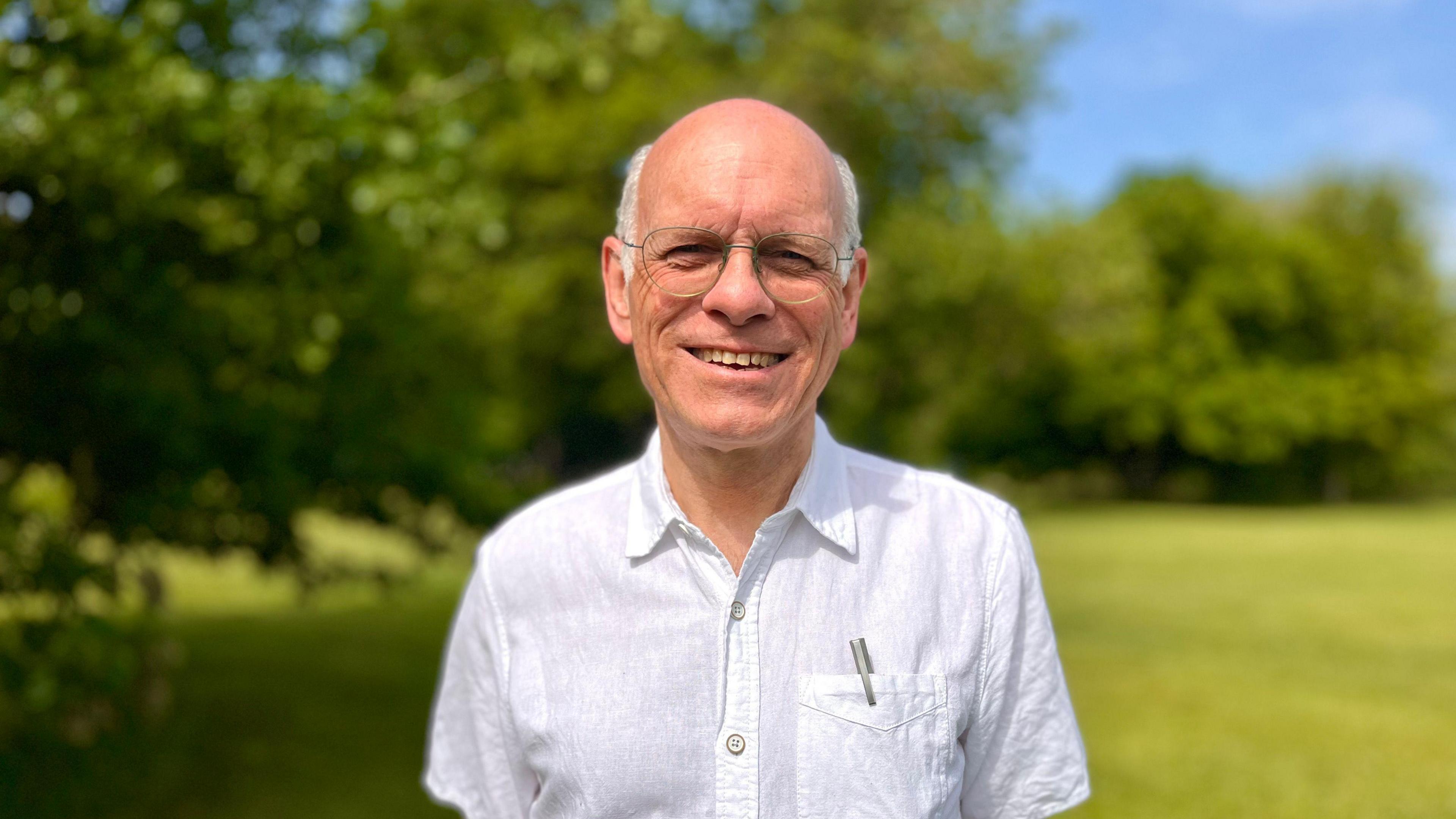 A smiling man faces the camera. He wears glasses, a white shirt with a pocket. A silver coloured pen is visible in the pocket. There is a blurred background of trees and a field
