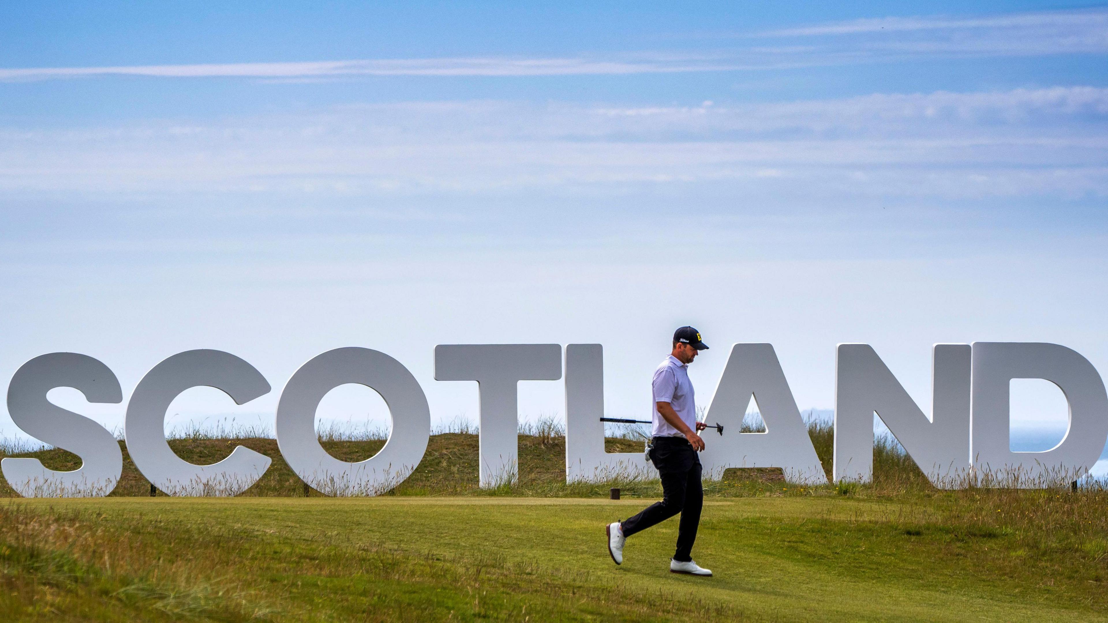 A lone golfer, photographed side-on, walks along the fairway past a Hollywood-style Scotland sign. A blue sky is visible in the background.