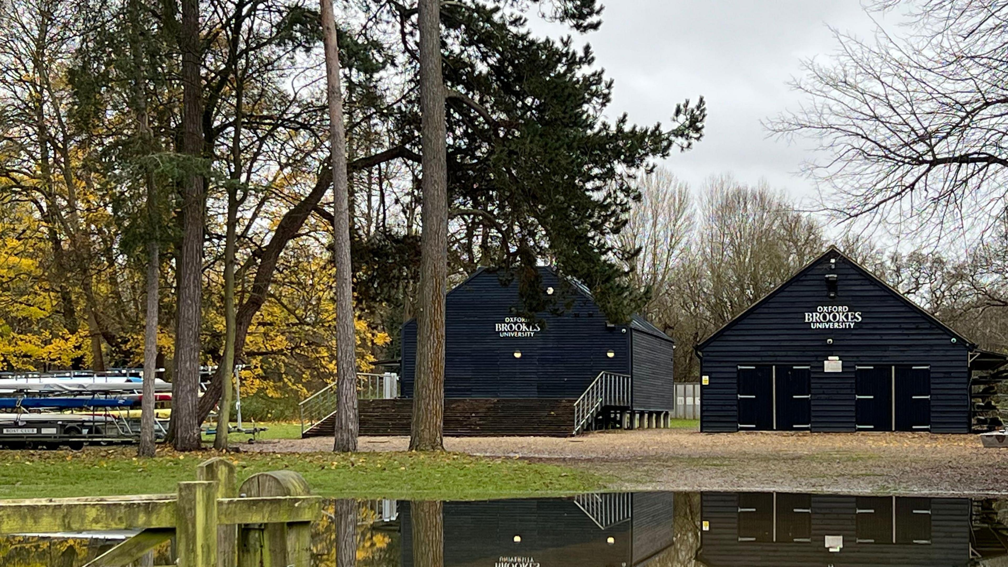 There are two wooden boat houses, painted black. On the front of them they have lettering saying Oxford Brookes University. Rowing boats are stacked in storage to the left of the boat houses. They are situated in a wooded, rural landscape and there are large puddles of water around.