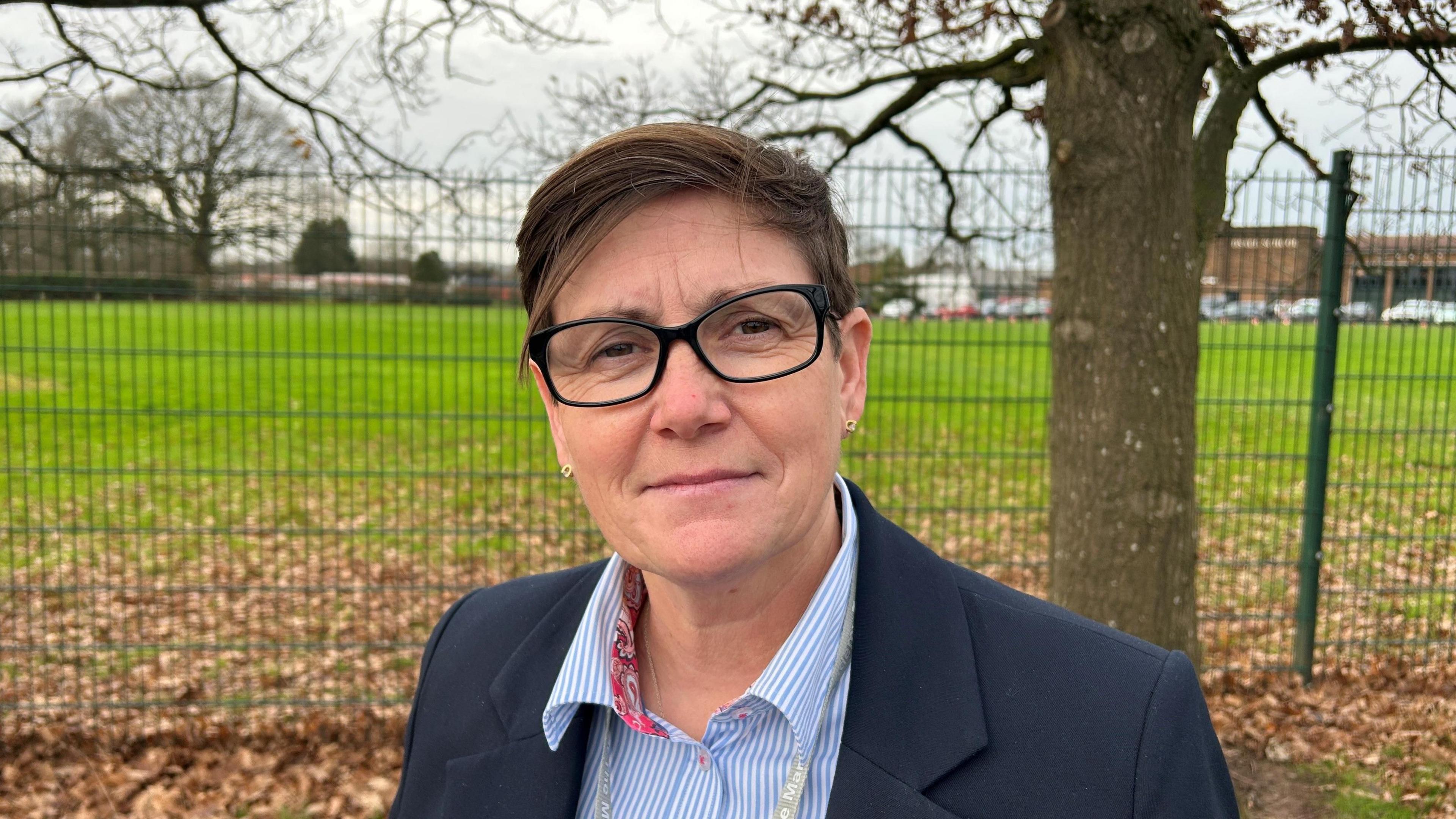 A woman with short brown hair and black framed glasses in a blue jacket and blouse standing in front of a fence with a leaf-covered field behind it