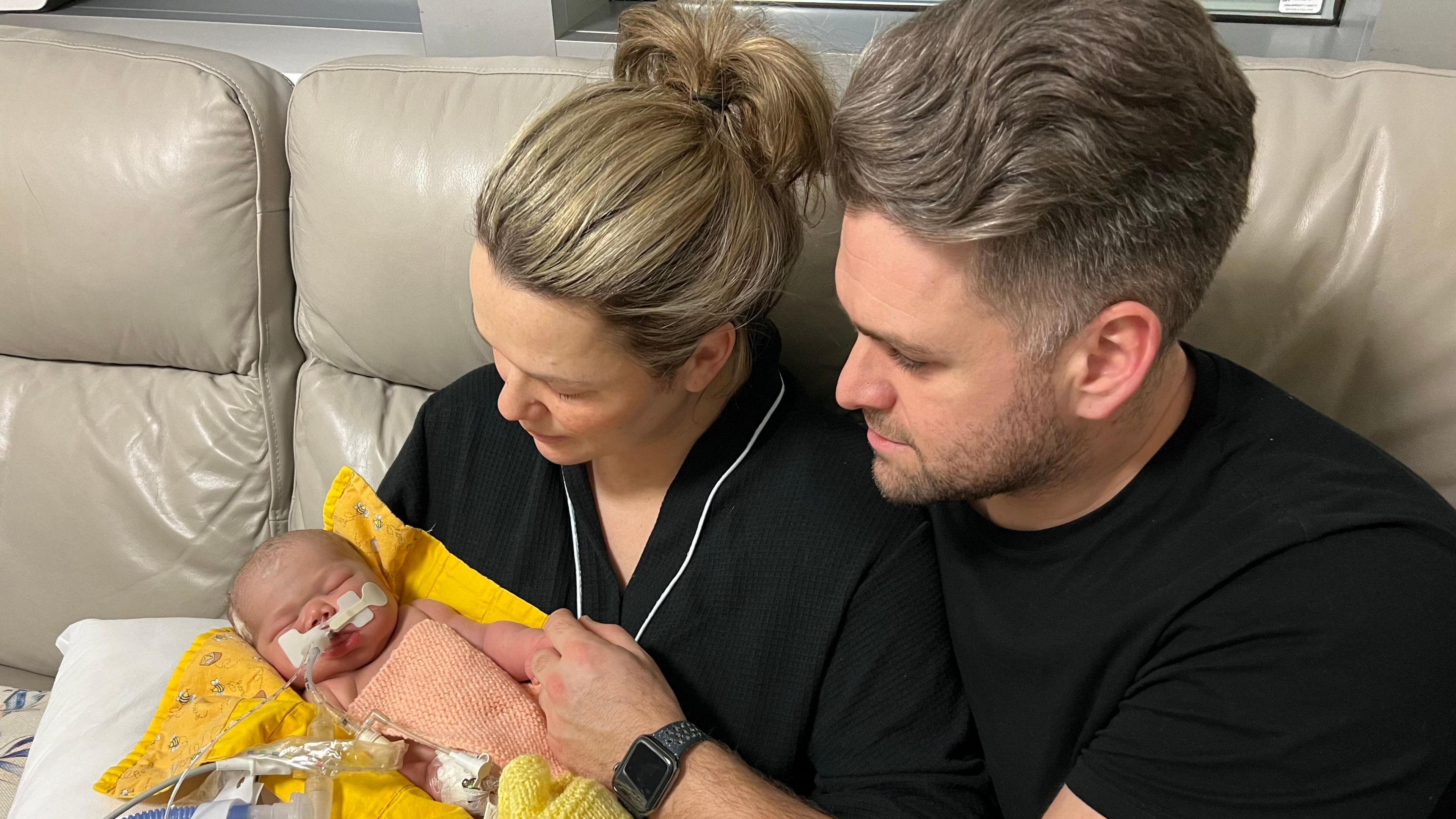 Bryony and Daniel Russo, both wearing black, sit on a cream sofa while cradling Emmy, who has tubes attached to her and is lying on a yellow blanket.