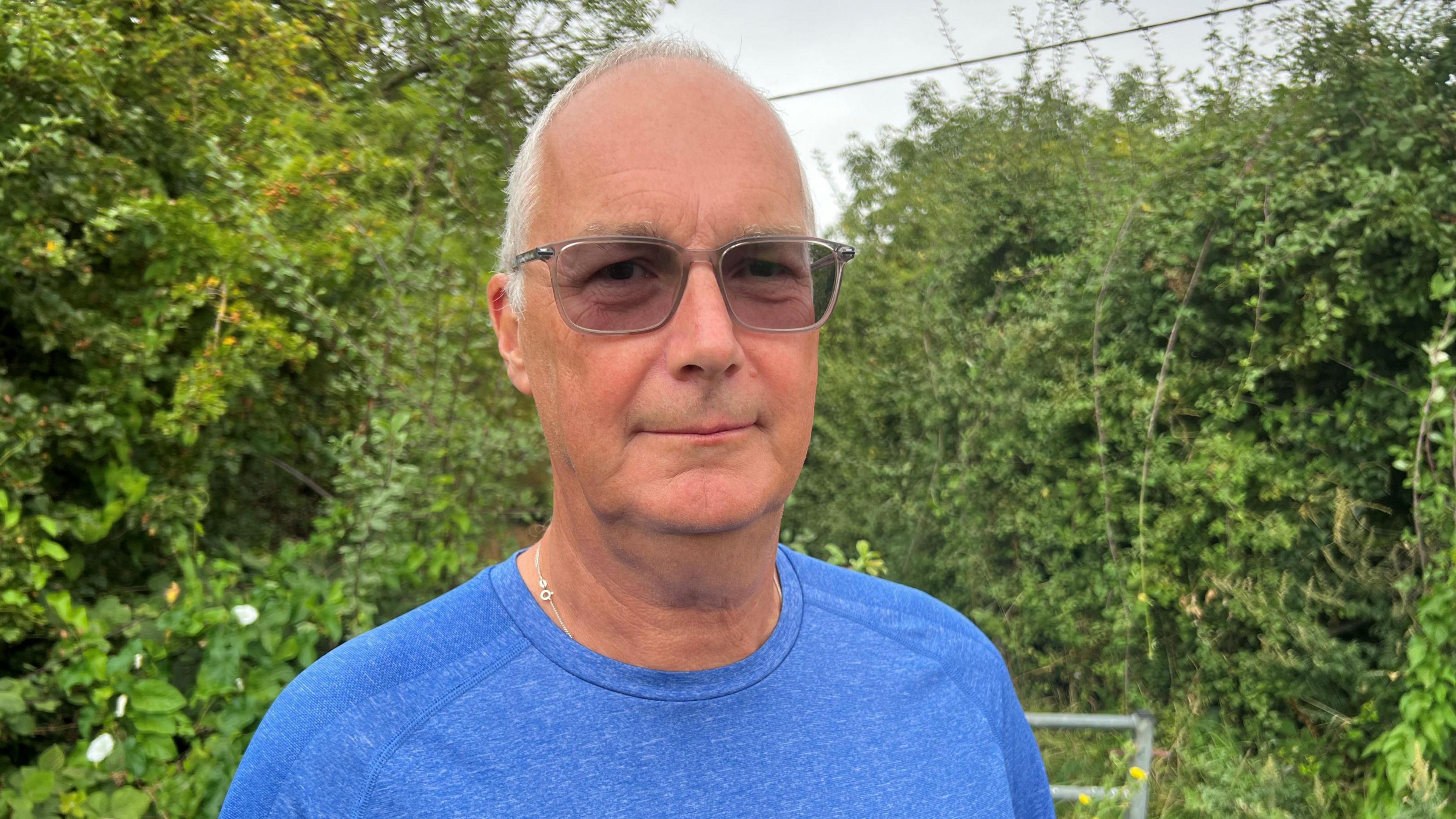 Mark Kelby - a man with thin grey hair and tinted glasses - looks straight at the camera. He is wearing a blue T-shirt and is standing in front of an area of hedge and trees. 