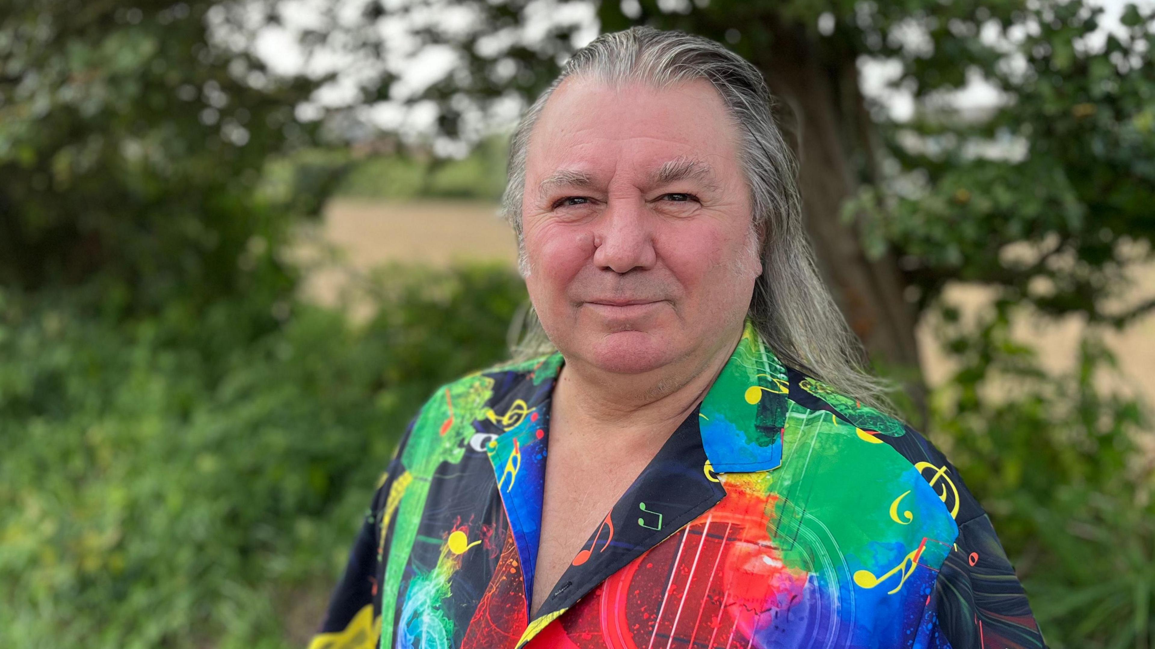 Mark Guillou in the outdoors, standing in front of a tree in a colourful shirt