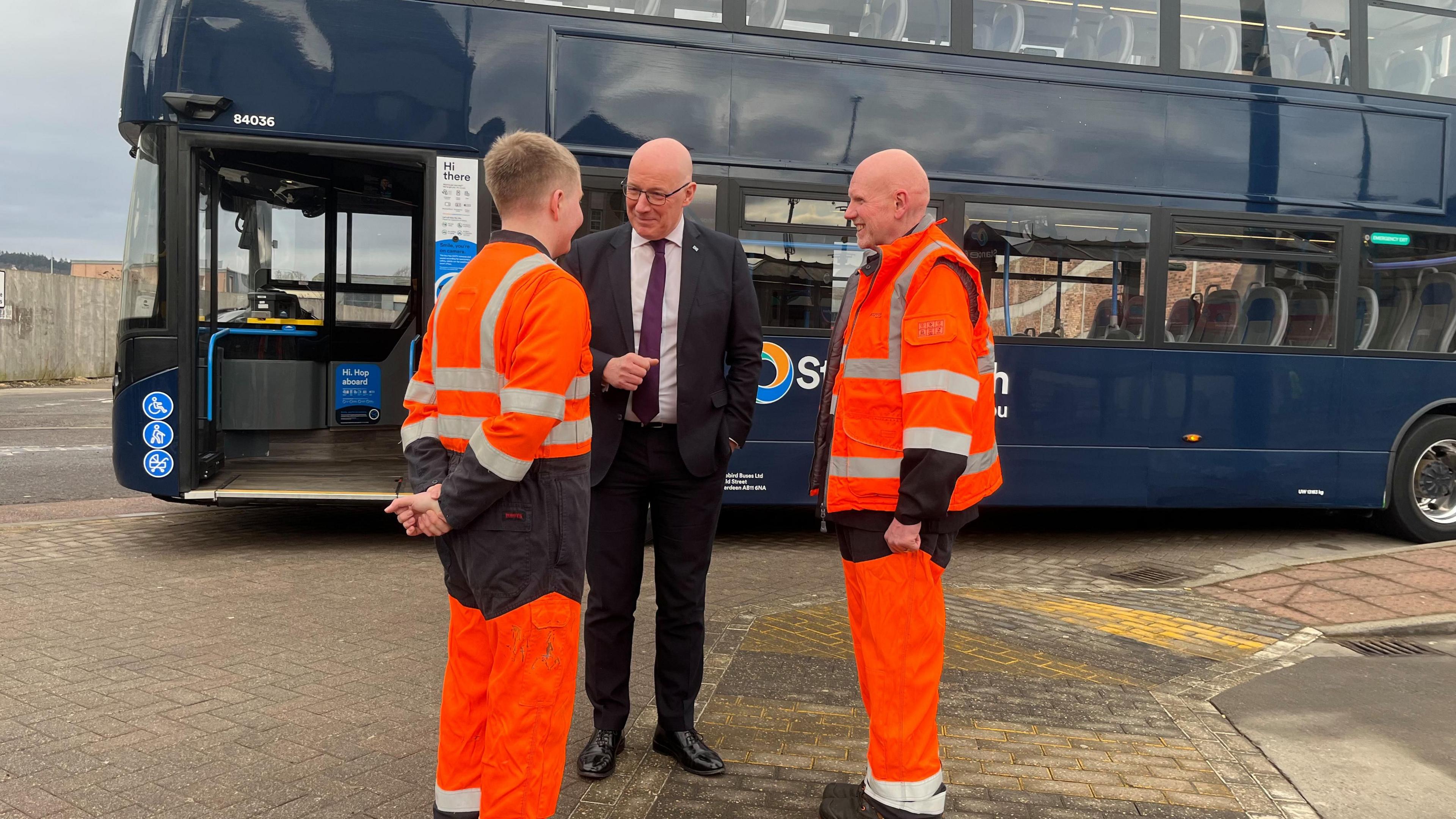 Tha Iain Swinney na sheasamh a' bruidhinn ri dithis fhireannach a th' air gabh taobh dheth. Tha esan ann an deise agus tha an dithis eile ann an deisichean h-vis orains. Tha bus dorch gorm air an cùlaibh. Tha iad a' gaireachdainn.