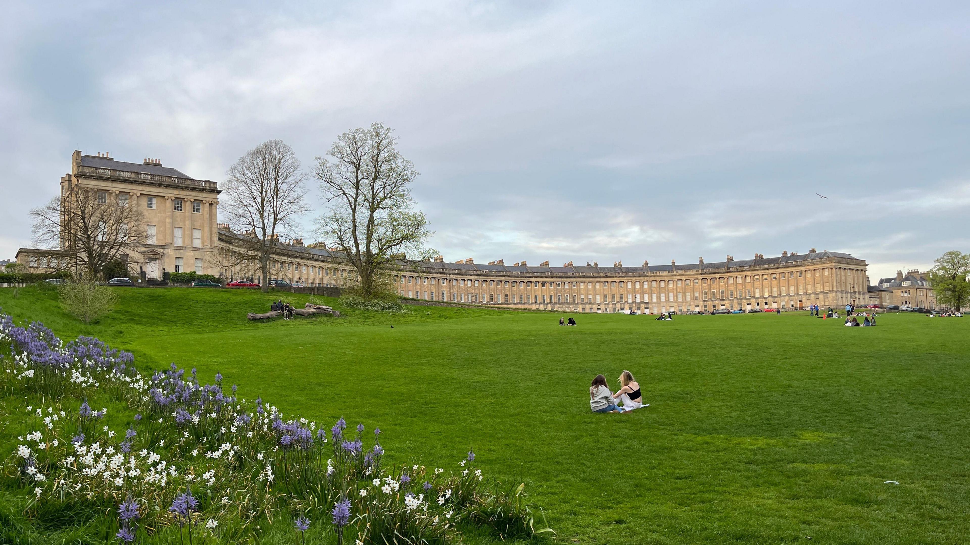 An image of the Royal Crescent lawn where a handful of people are sitting and enjoying a picnic. The iconic row of Grade I terrace houses are in the background.