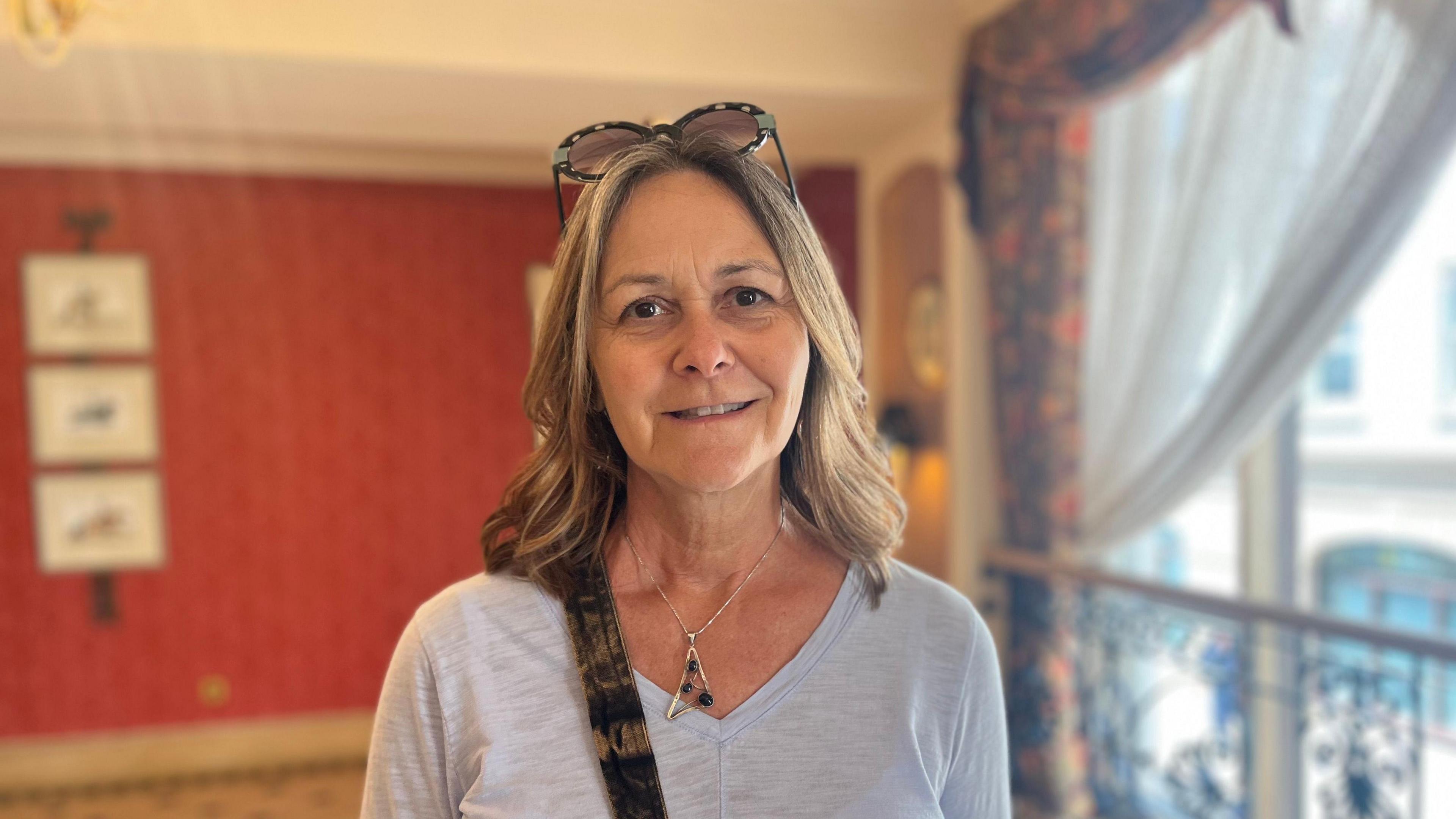 Kim Cisna is standing in a hotel reception room with red walls and a large window. She is wearing sunglasses on her head, with a grey top. She is smiling into the camera.