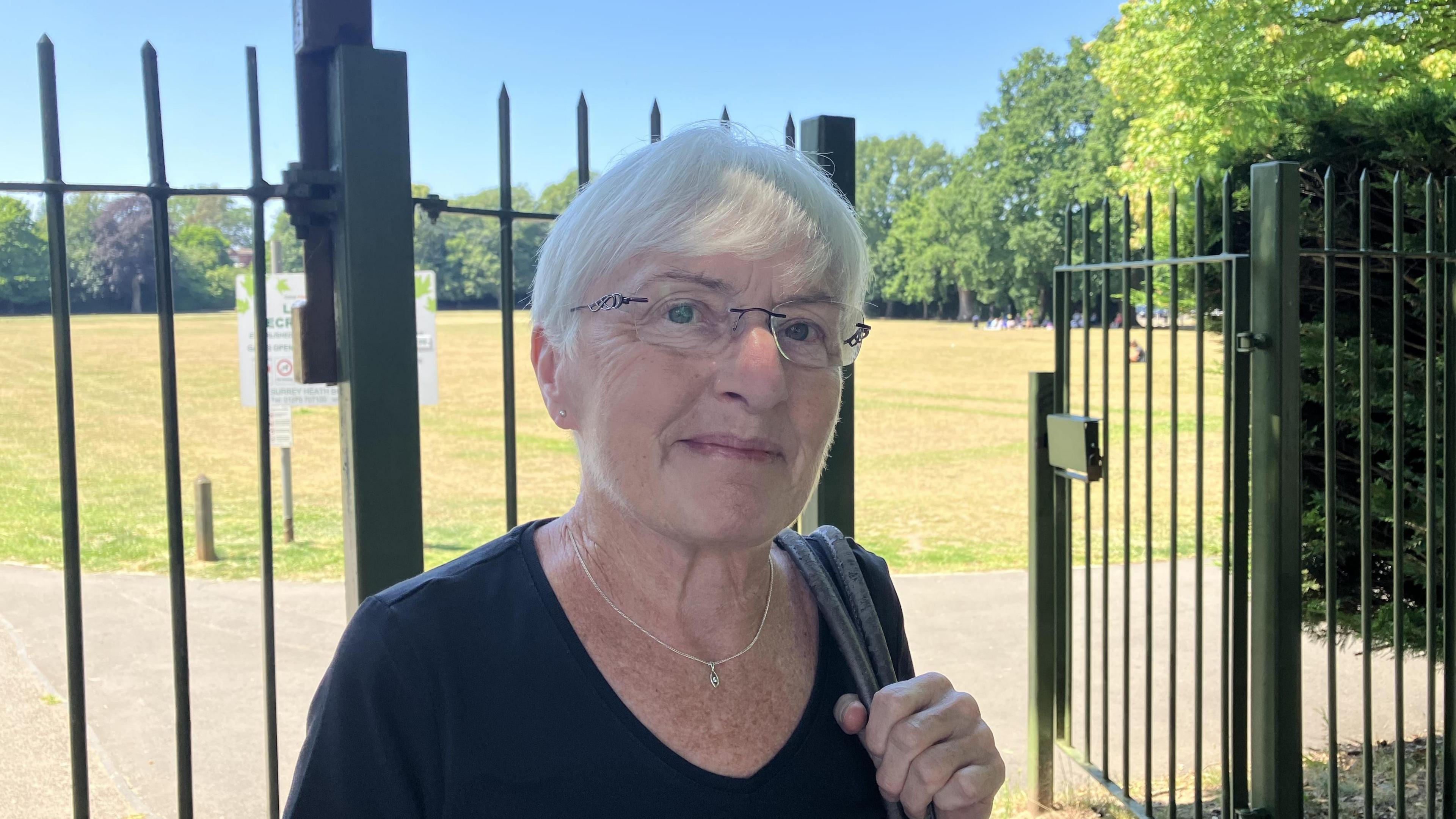 A woman with white hair and a bag over her shoulder. The park behind her is basked in sunlight.