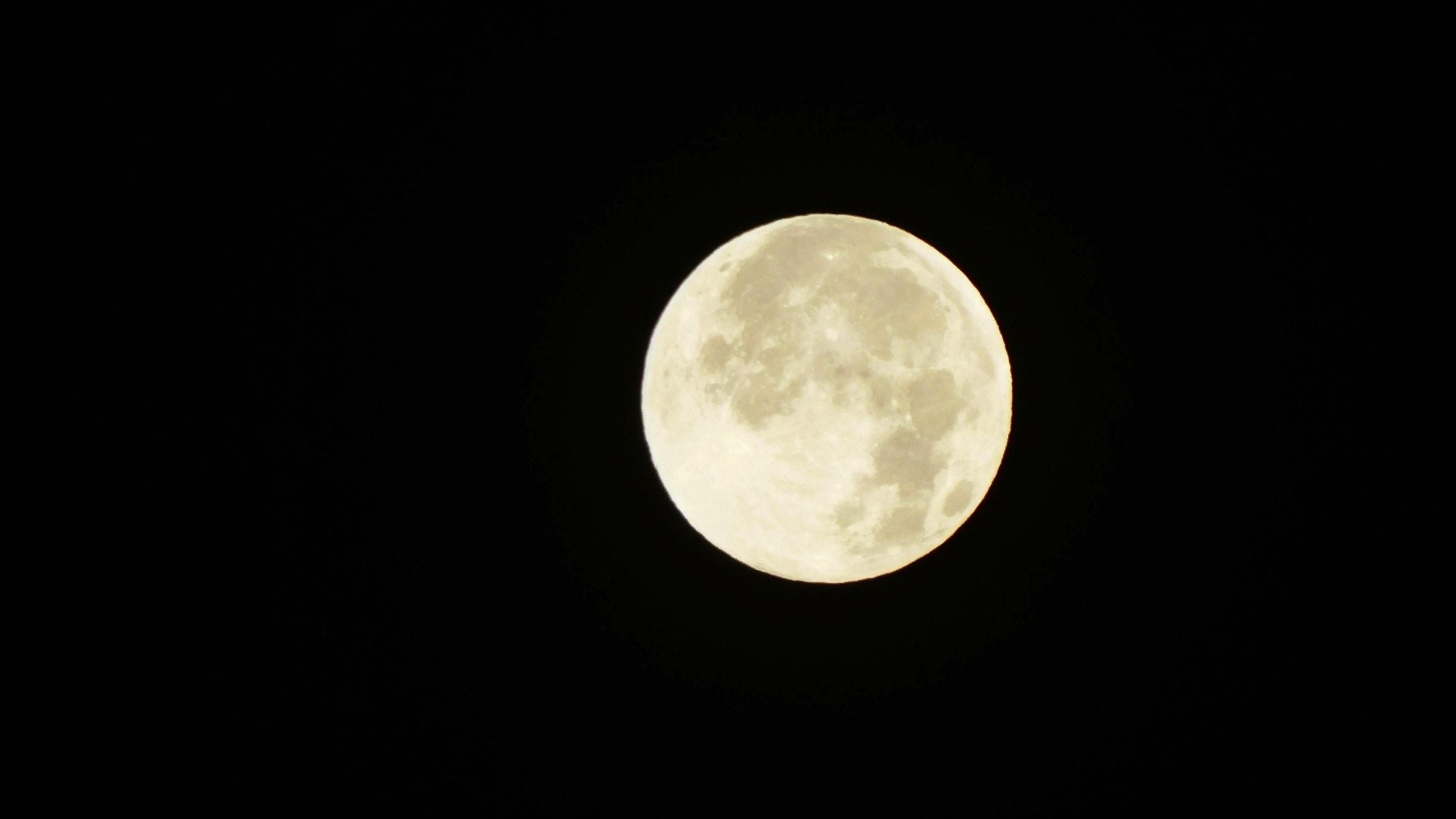 A picture of a bright white circular Moon with darker beige patches dotted across. The moon is a stark contrast to the pitch dark background.