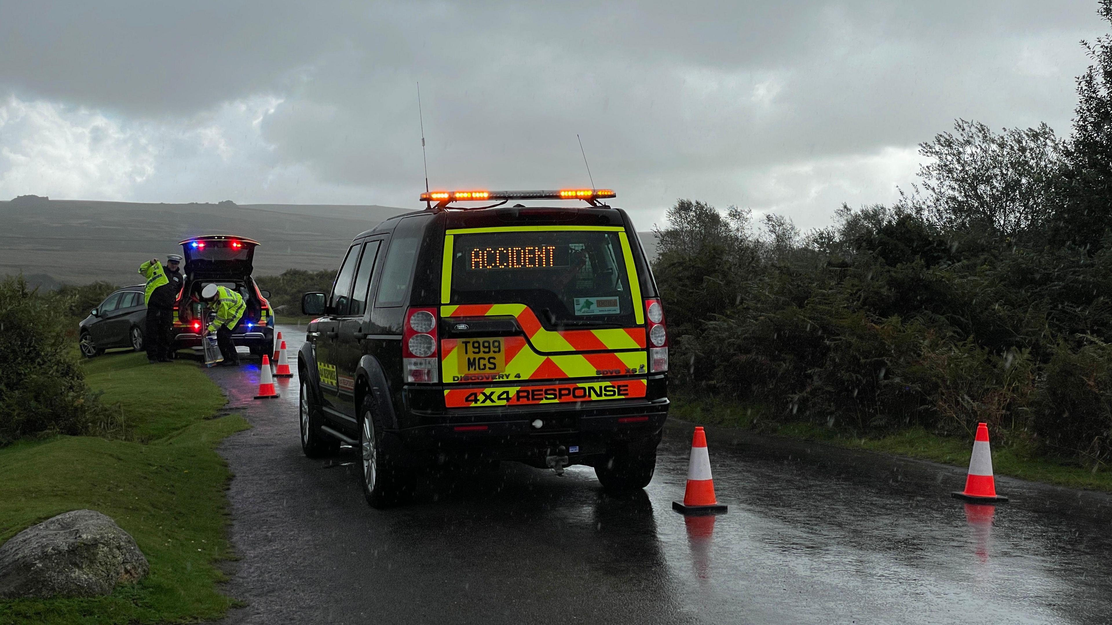 A 4x4 vehicle with high-viz markings on it and the word 'ACCIDENT' in the rear window in LED lights. It is on a narrow moorland road with traffic cones behind it and a police car in front.
