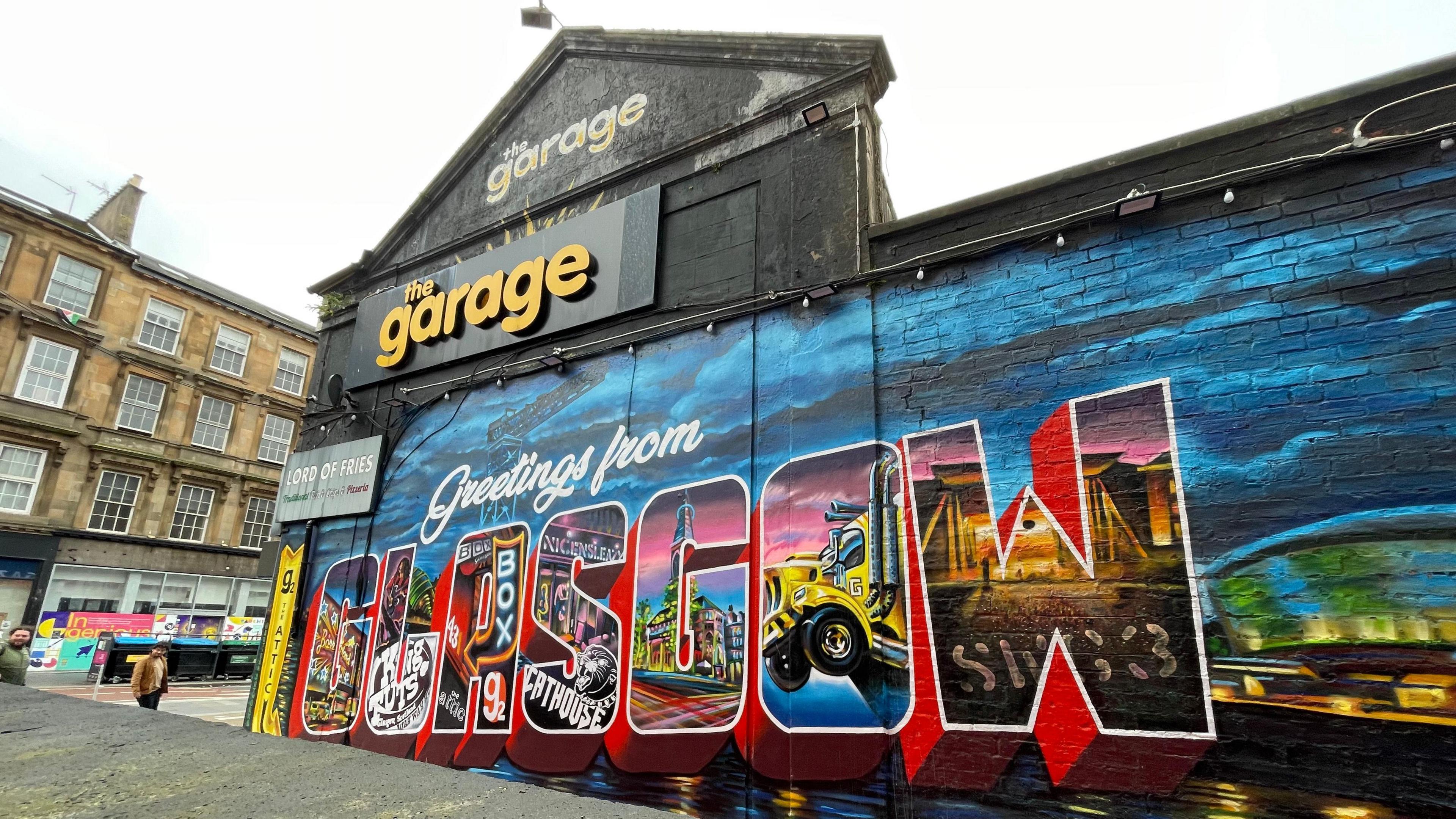 An outside shot of the Garage in Glasgow with a mural reading 'Greetings from Glasgow' on the side.
