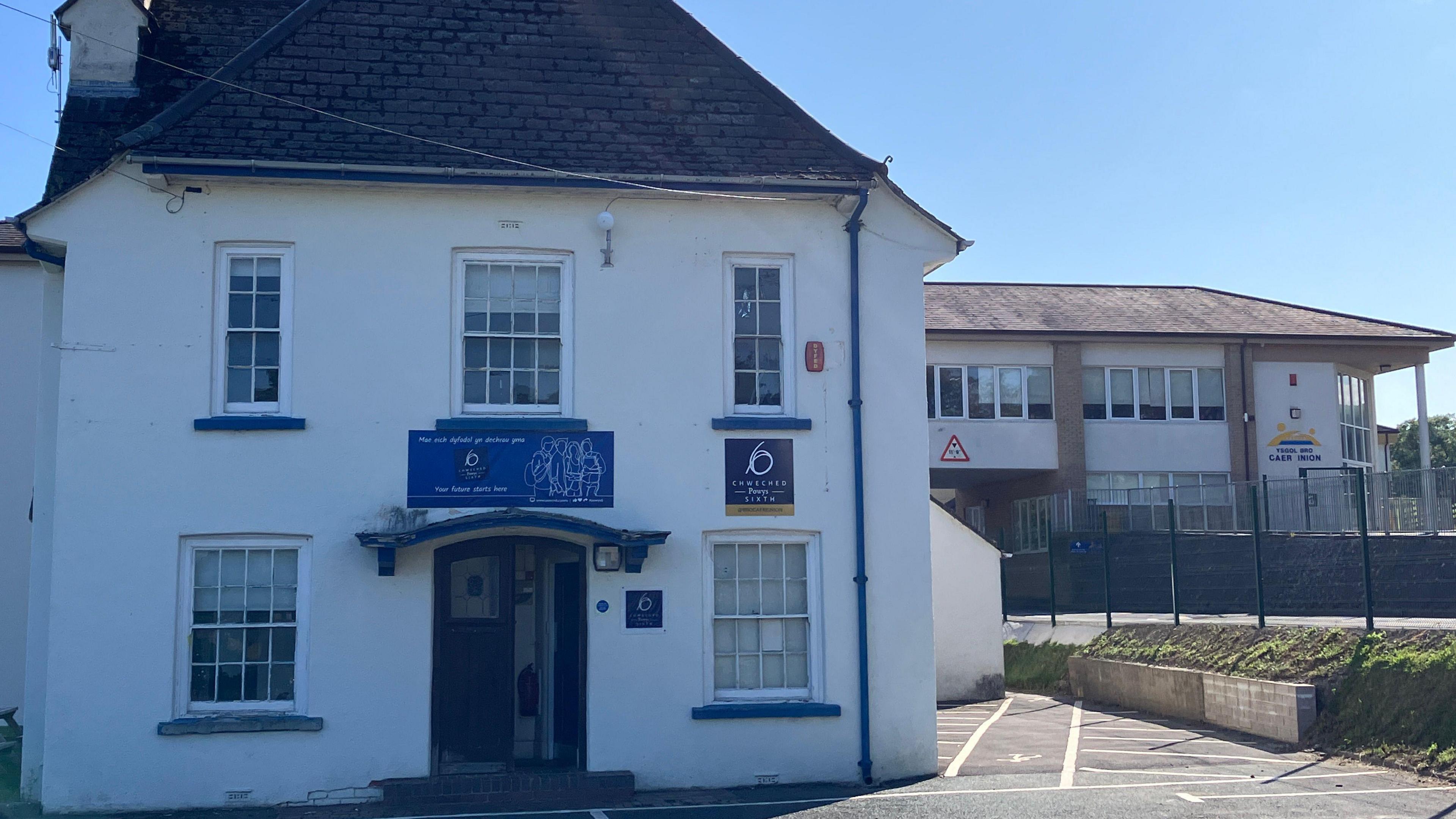 The entrance of Ysgol Bro Caerinion with a door leading to a white school house. There is a road to the right that runs underneath a second floor bridge and a school yard can be seen on the right hand side. 