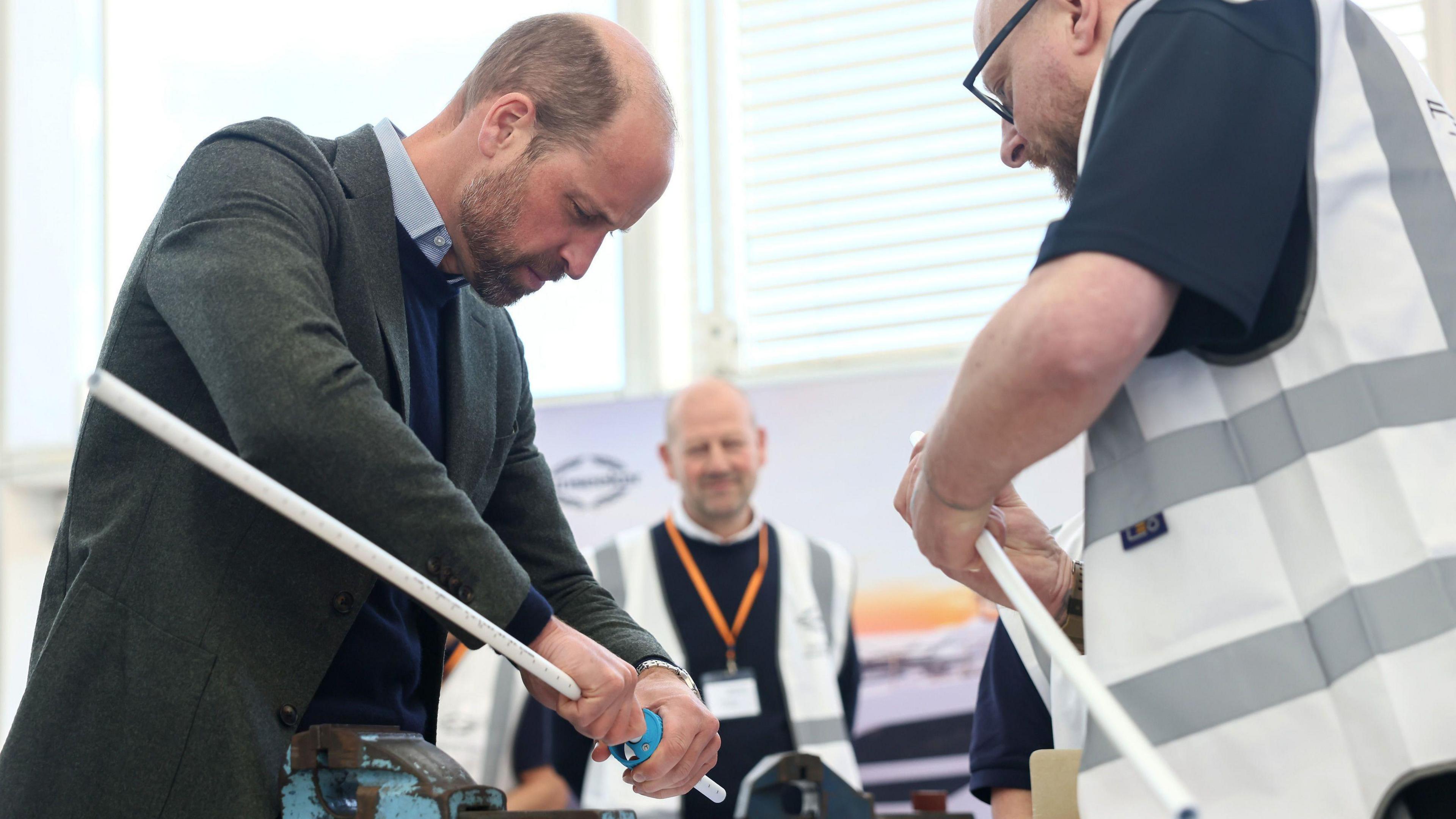 Prince William holding a long thin white pipe in his right hand and cutting it with a small blue pipe cutter held in his left hand. He is being watched by two men wearing white hi-viz vests.