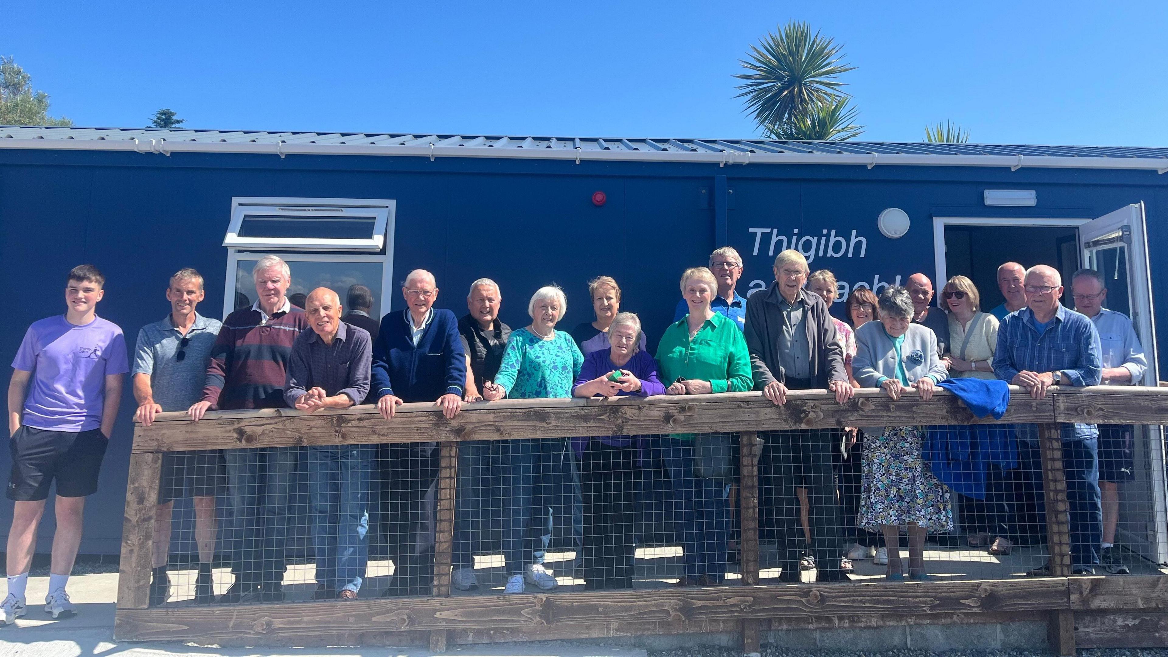 Dealbh de chuid de na daoine a bhios a' gabhail pàirt sa chluba gach Diciadain agus iad nan seasamh taobh a-muigh an togalaIch a bhios iad a' cleachdadh airson coinneachadh. Tha là math ann 's an adhar gorm. Tha gach neach a' coimhead uabhasach toilichte.
