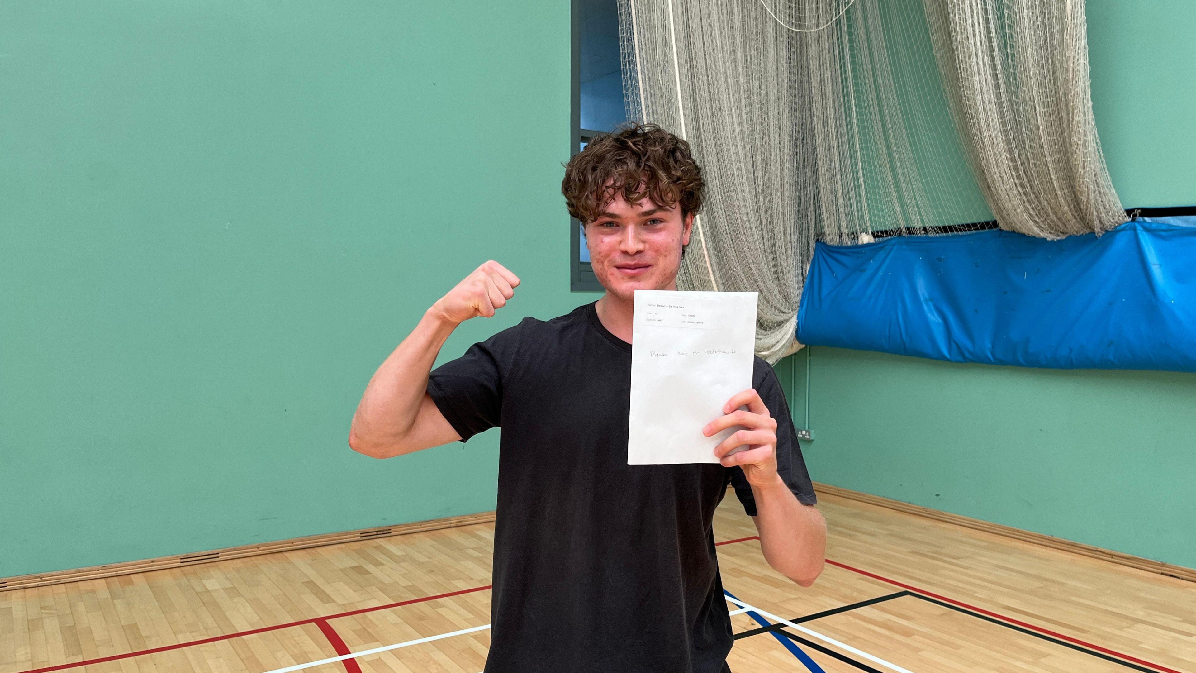 Isaac wearing a black T-shirt holding his results in his hand- standing inside the school sports hall