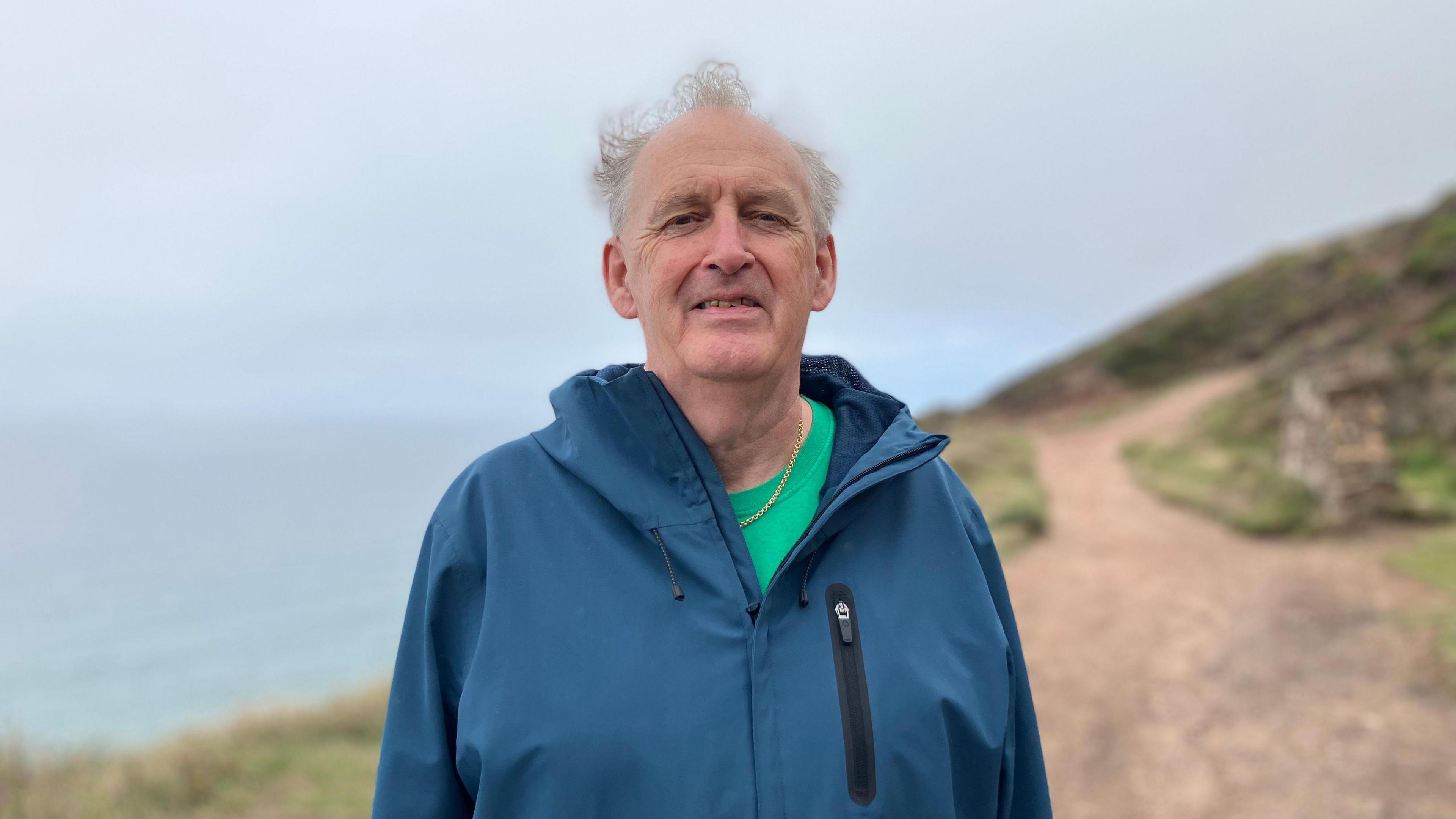 Pat Dunne smiling at the camera. He is wearing a green t-shirt and a blue coat. He is stood on a coastal path with the sea behind him.