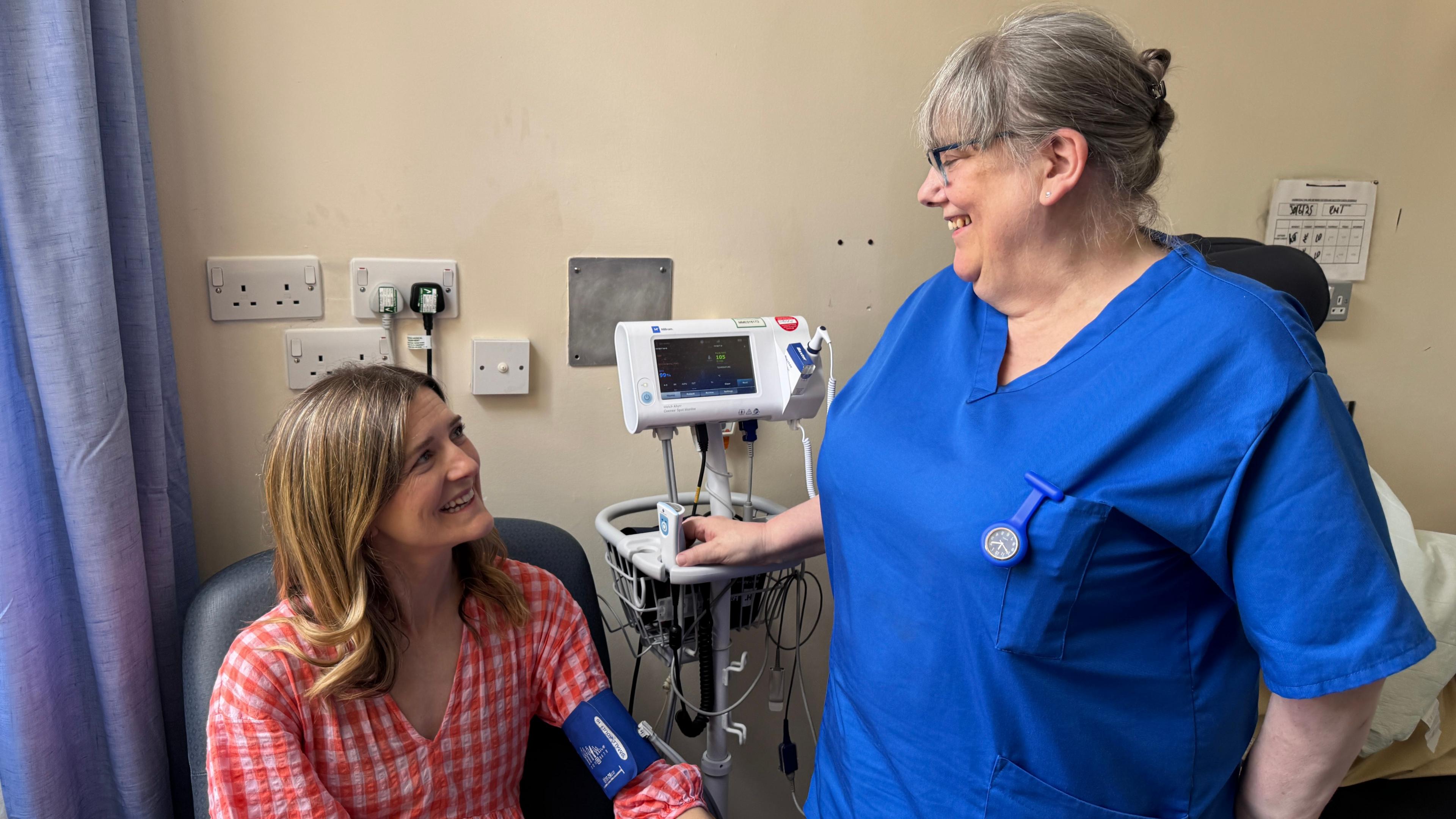 Patient, Hannah Robinson, wearing a red and white checked dress sitting in a hospital room smiling at a nurse standing in front of her, wearing a blue tunic and taking her blood pressure
