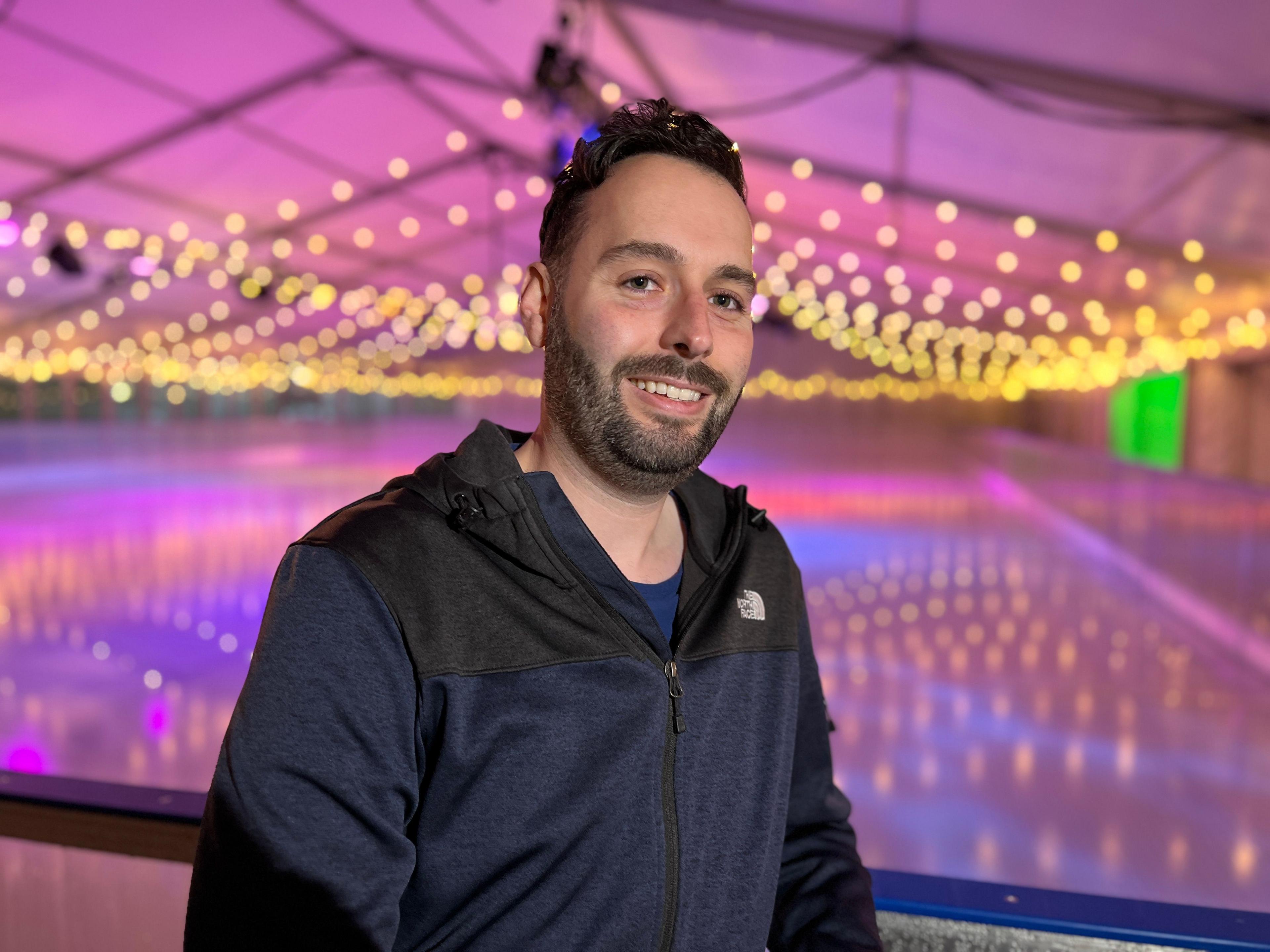 Smiling man standing sideways in front of an indoor ice rink. The roof is lit up with bright lights. The man, who has short brown hair with a slight wave, is wearing a zipped North Face top which is black and blue. He has a lighter blue t-shirt underneath. 