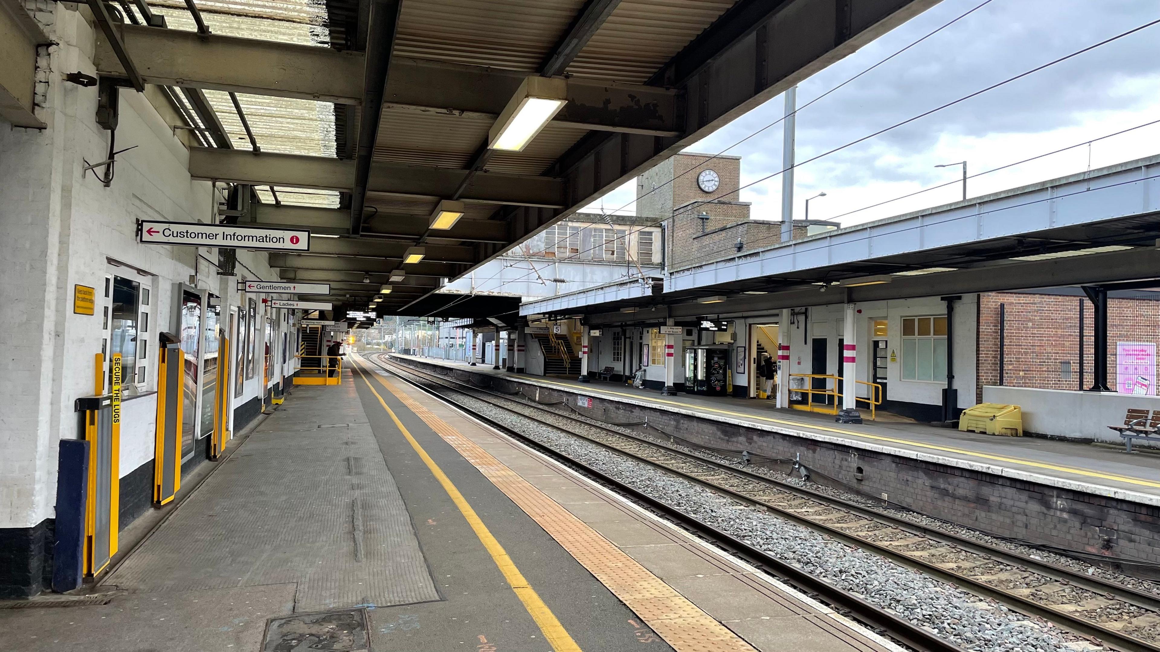 A look along a near-deserted platform at Luton station, with tracks sweeping away into the distance, from bottom right into the middle-left of the image. The station's 1930s clock tower is just visible in the background, as is a footbridge over the tracks.