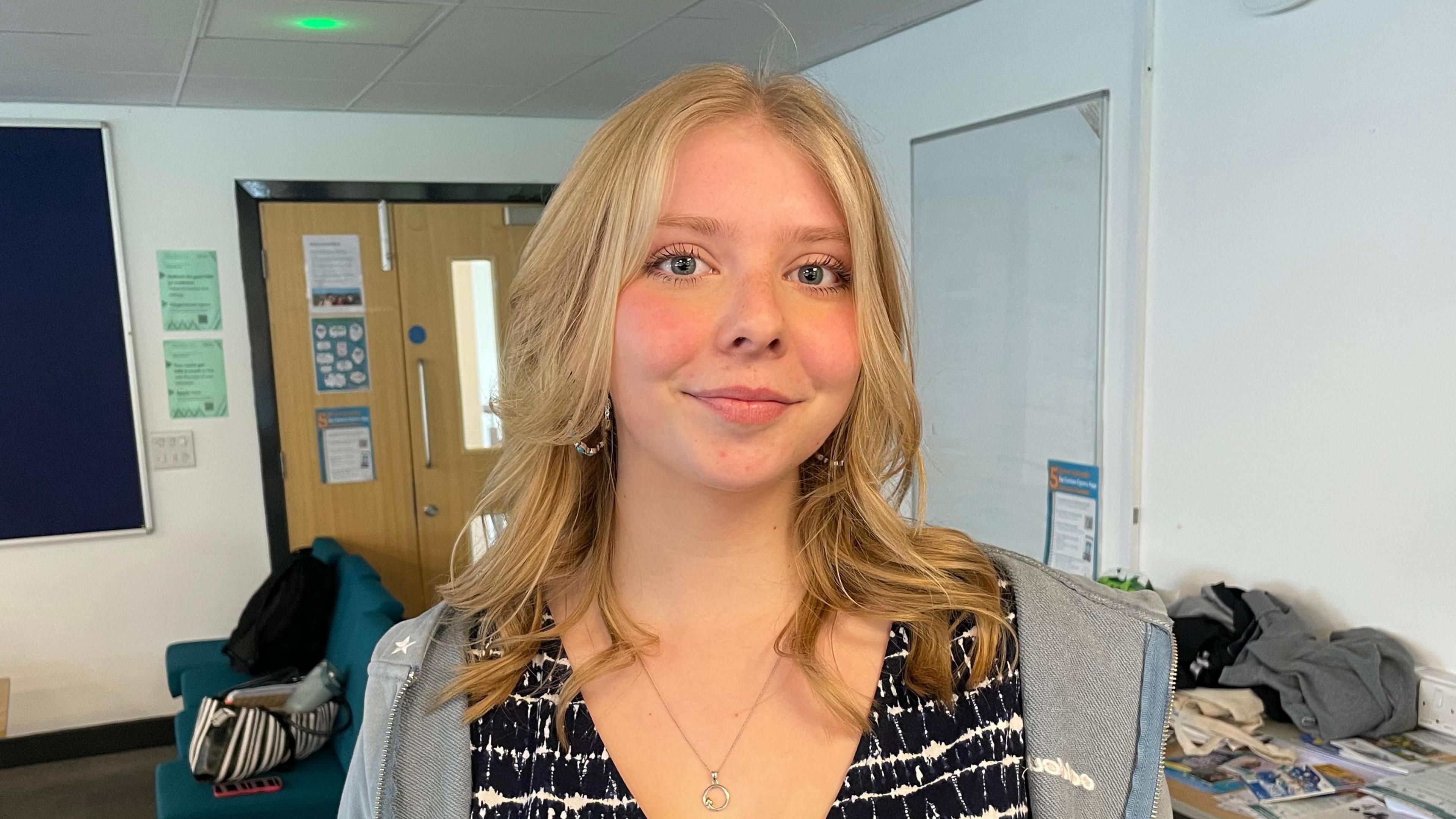 Merch gyda gwallt melyn i'w hysgwyddau gyda top du a gwyn a hoodie llwyd a mwclus yn sefyll mewn lolfa chweched dosbarth mewn ysgol