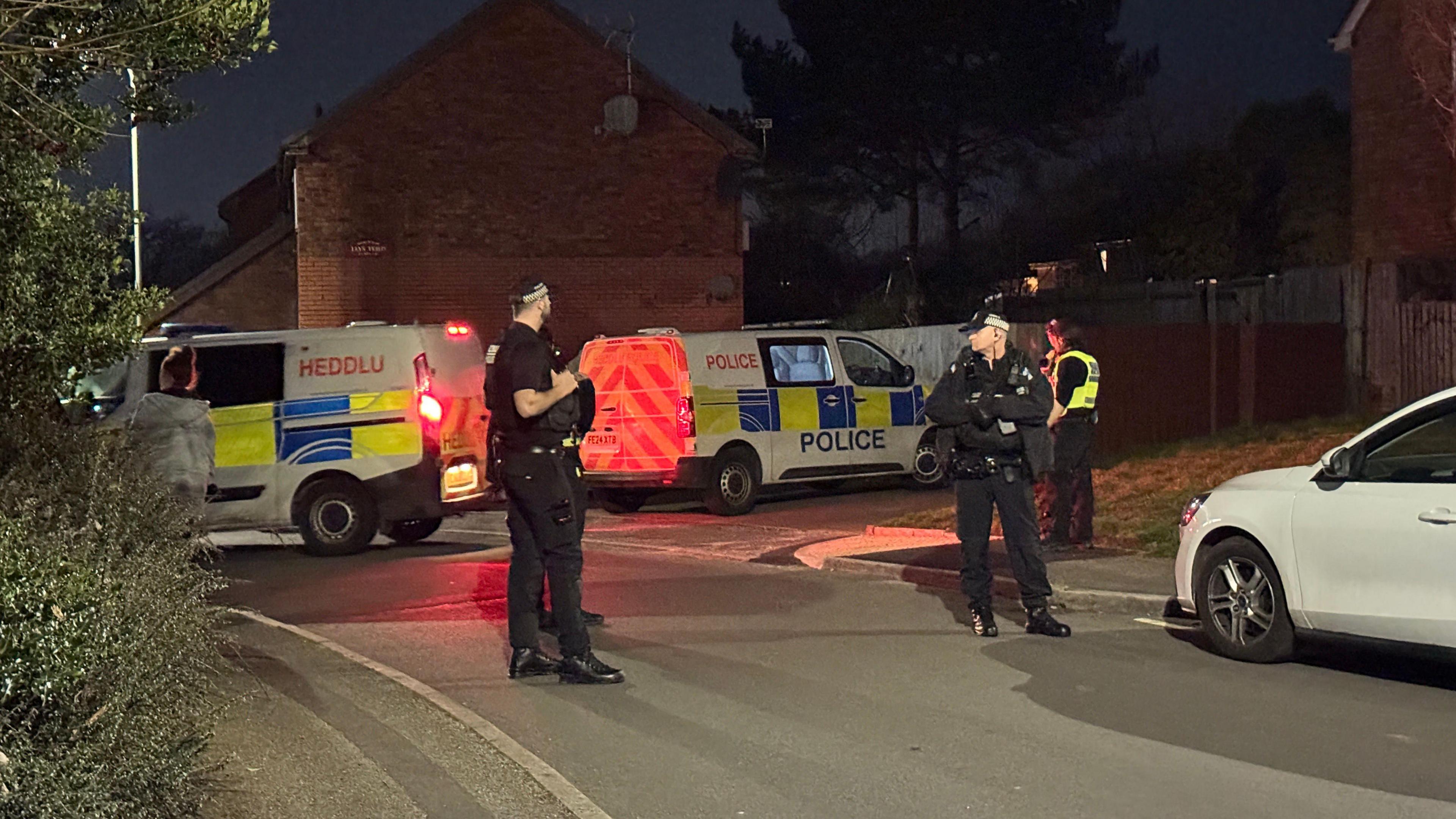 Three police officers can be seen standing on the street, with two police vans behind them. It is dark, and one van has its brake lights on. Two police officers are wearing a black uniform with black caps on. A third is slightly in the background facing away, wearing a yellow fluorescent vest. 