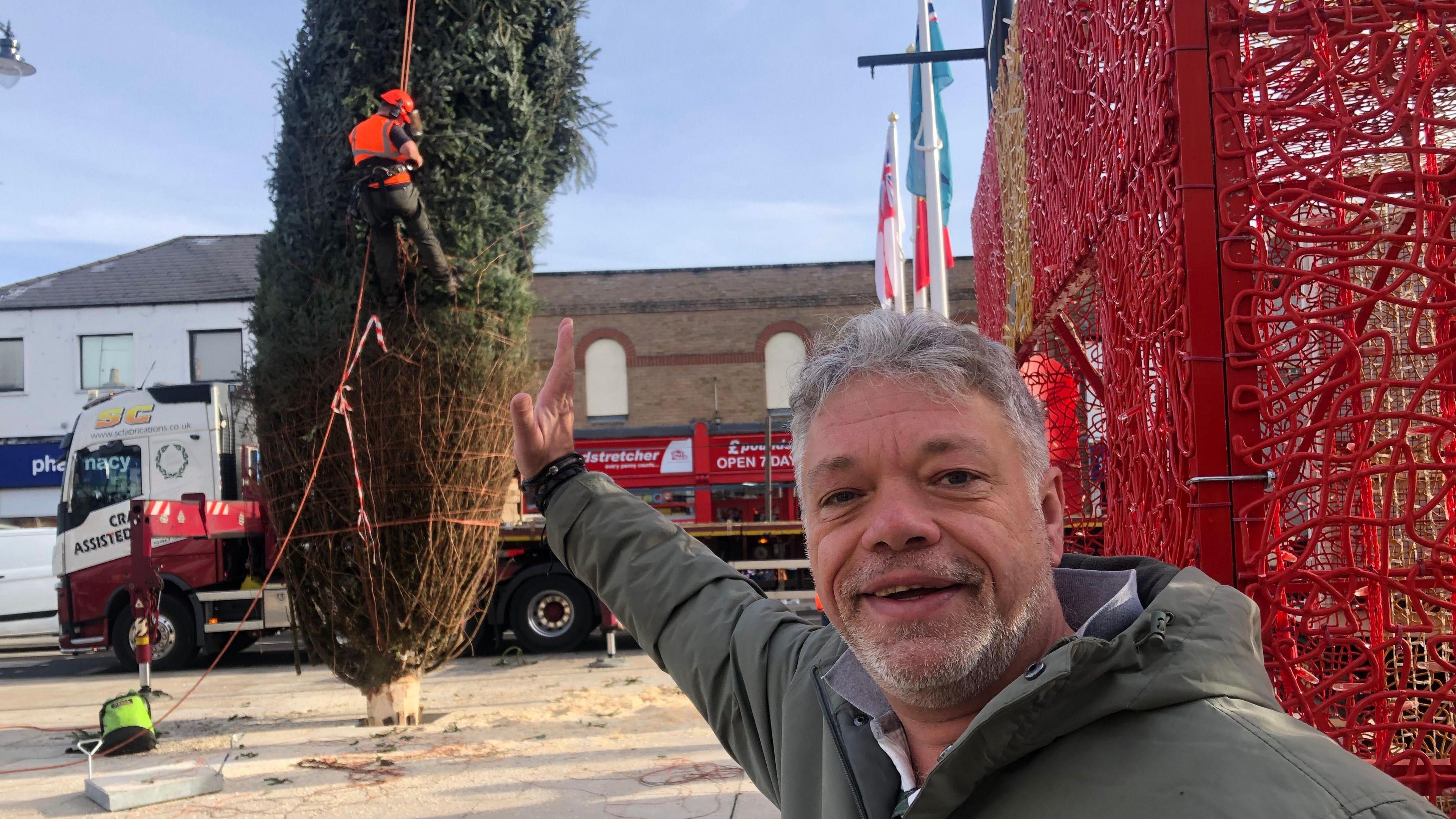 Paul Hicks has short grey hair with a grey beard and moustache and appears to be using his right arm to keep the leaning tree upright. He is wearing a grey overcoat with a white shirt underneath, with the collar up. Behind him is the leaning tree, with netting on it and a man in an orange hi-vis jacket and hard hat pulling the netting off