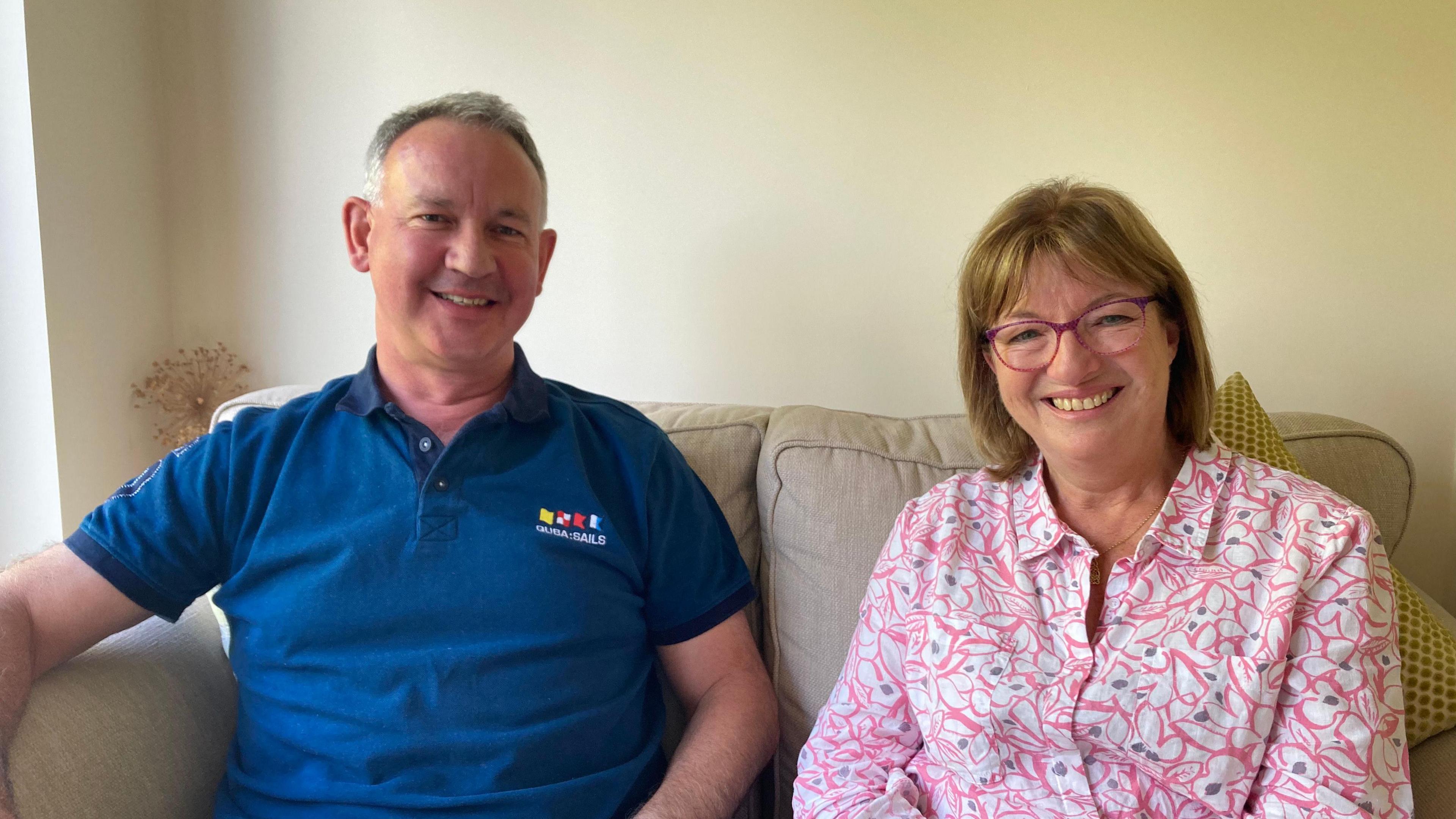 David Lowe and Hilary Lowe smile directly at the camera as they sit on a beige sofa, David is wearing a blue polo neck and Hilary a pink patterned shirt