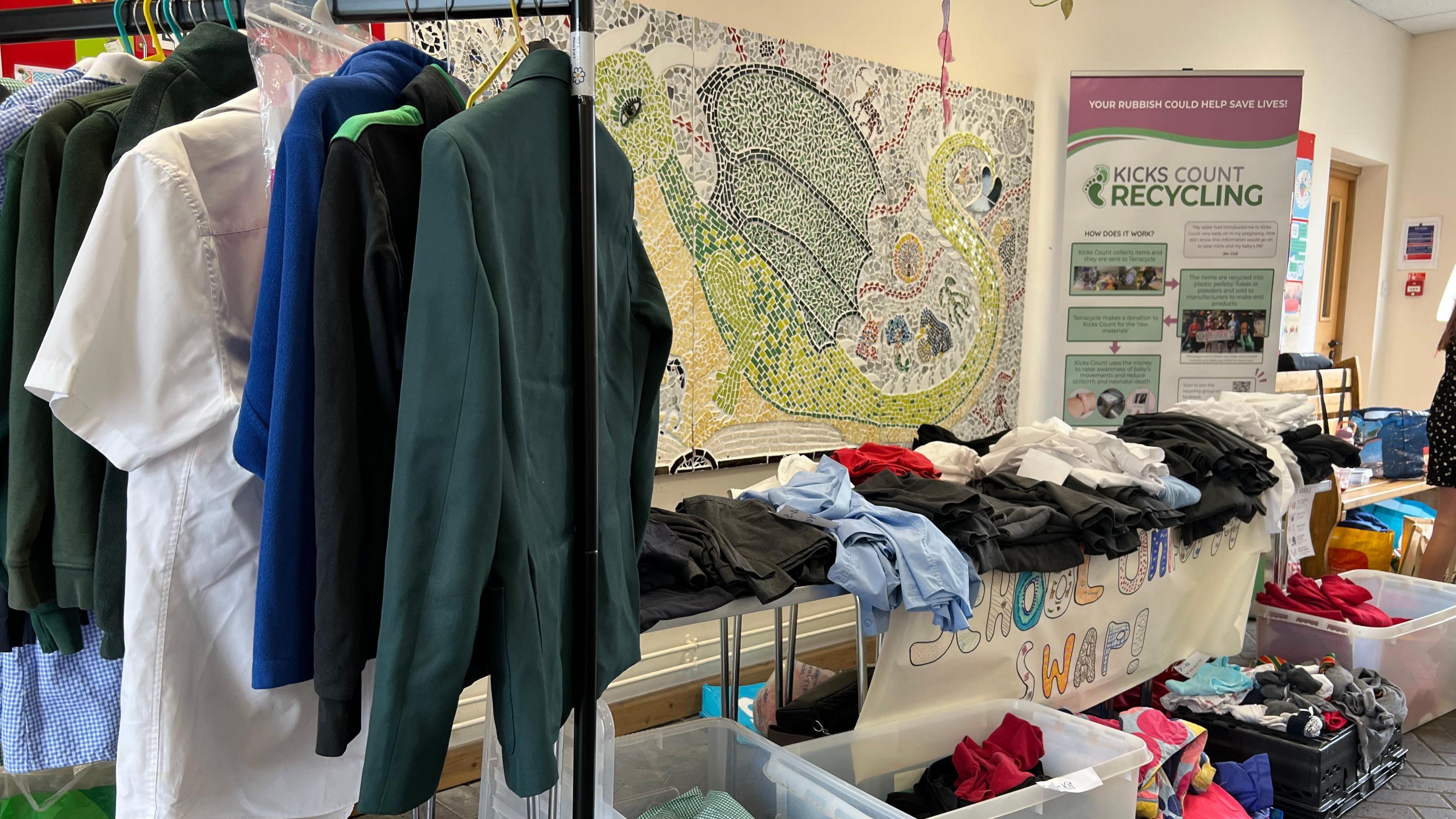 A view of school uniform items on a rail to the left, and on the table, a range of different coloured clothing on a table. Underneath the table are boxes filled with more items. 