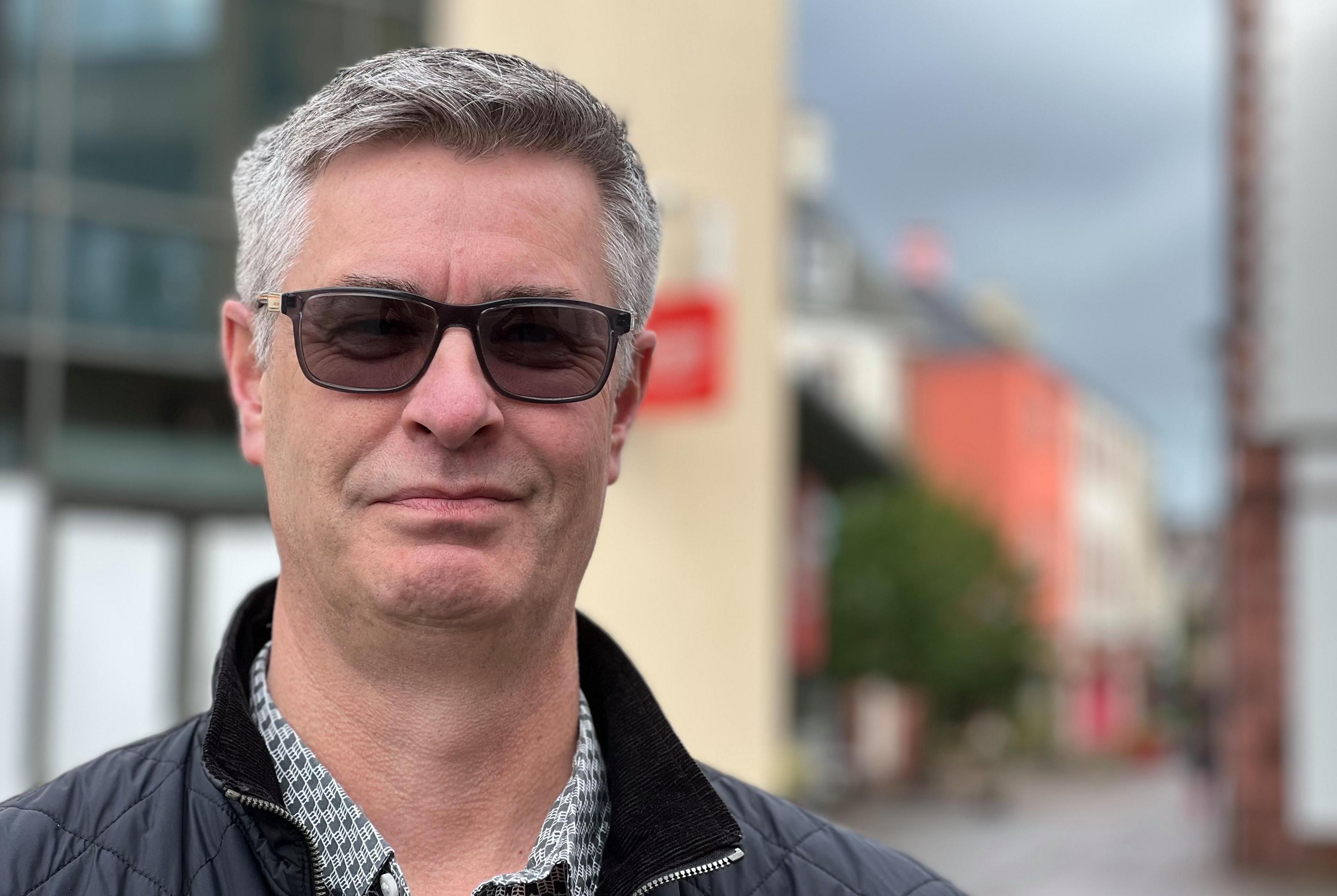 Darren Broad, chair of Penrith Business Improvement District, is standing in front of the entrance to New Squares in the town centre, wearing sunglasses and a black jacket.