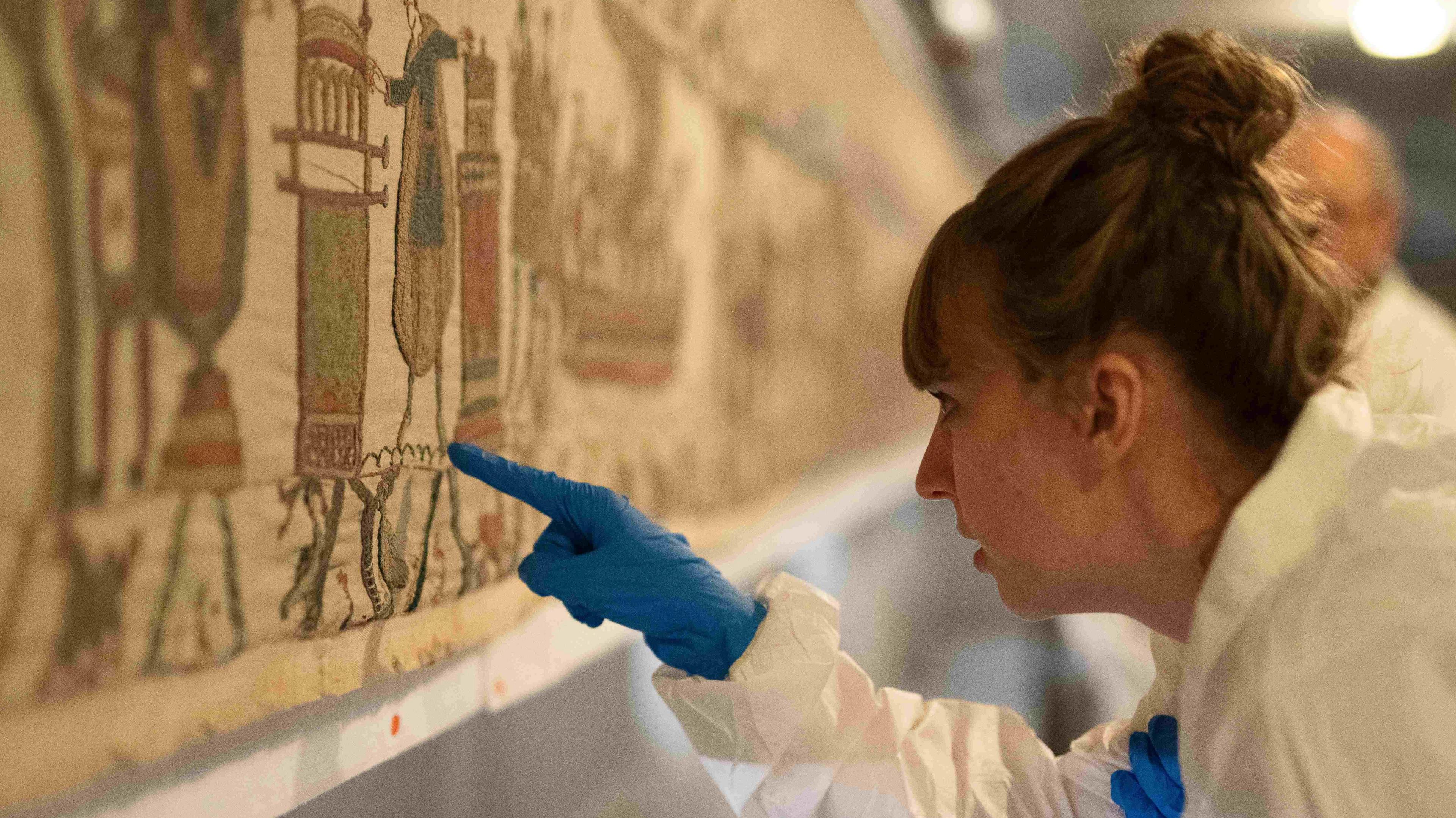 A woman in a sterile suit and gloves inspects the artwork with a pointed finger.