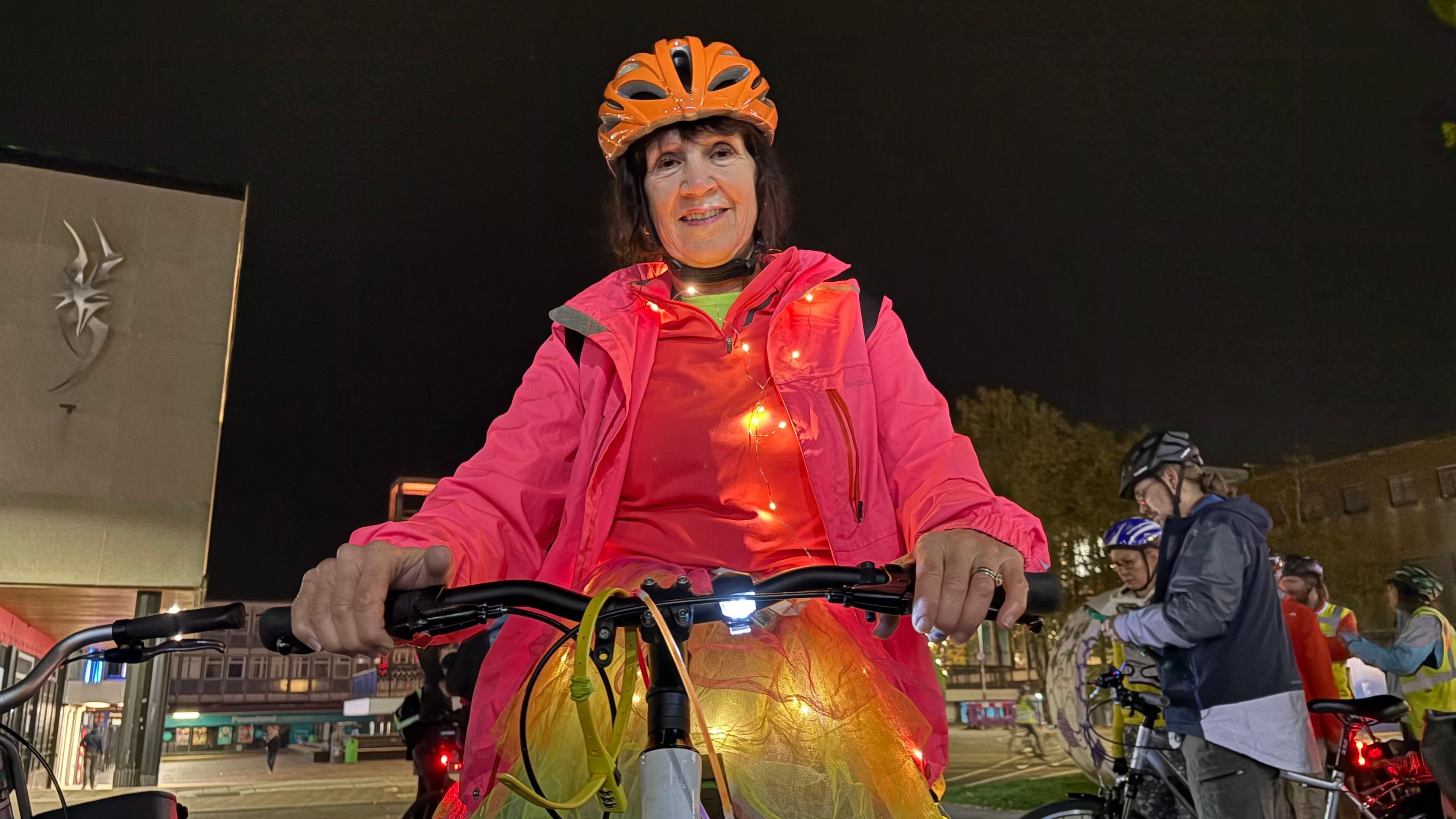 Lindsey Day wearing an orange cycle helmet, red sweater and pink jacket. She is riding a bike and has a string of fairy lights on top of her clothes. There are more cyclists in the background and there is a tall building to the left.