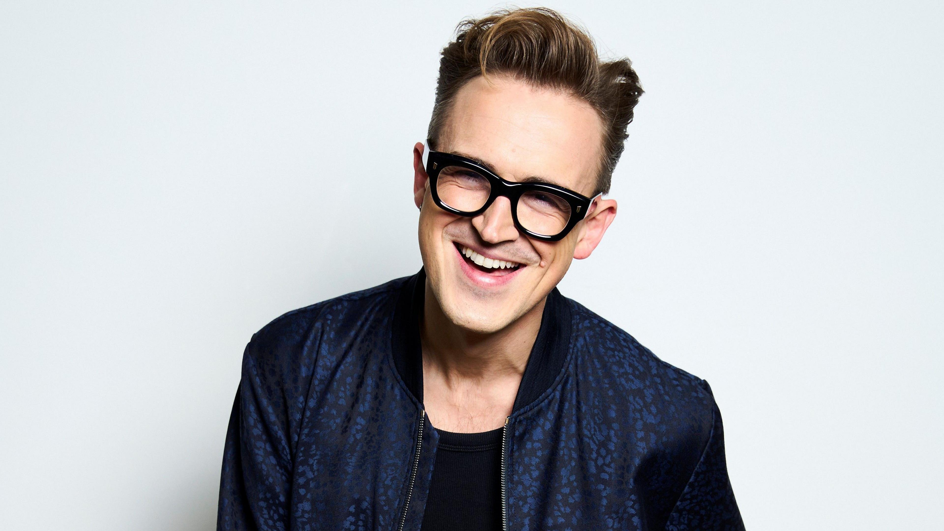 Tom Fletcher is standing in front of a white background. He is laughing and wearing thick-rimmed black glasses. His hair is swept up and back. He is wearing a plain black t-shirt beneath a patterned dark blue jacket.