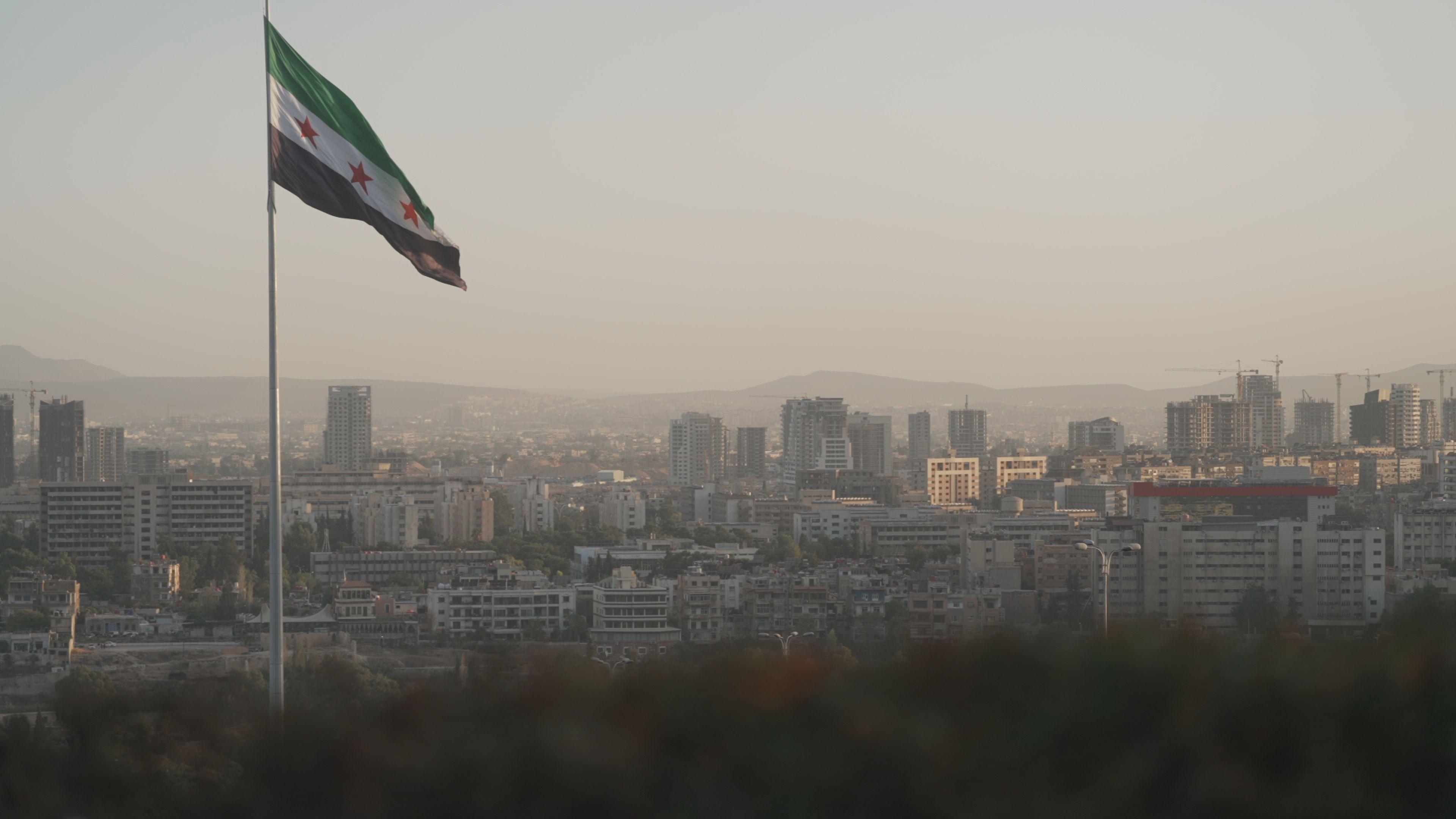 A cityscape surrounded by mountains in the distance is captured in a red dusk like light. A single flag pole has a large flag catching the wind. It has a green stripe at the top, a white stripe bearing three red stars, and a black stripe at the bottom.