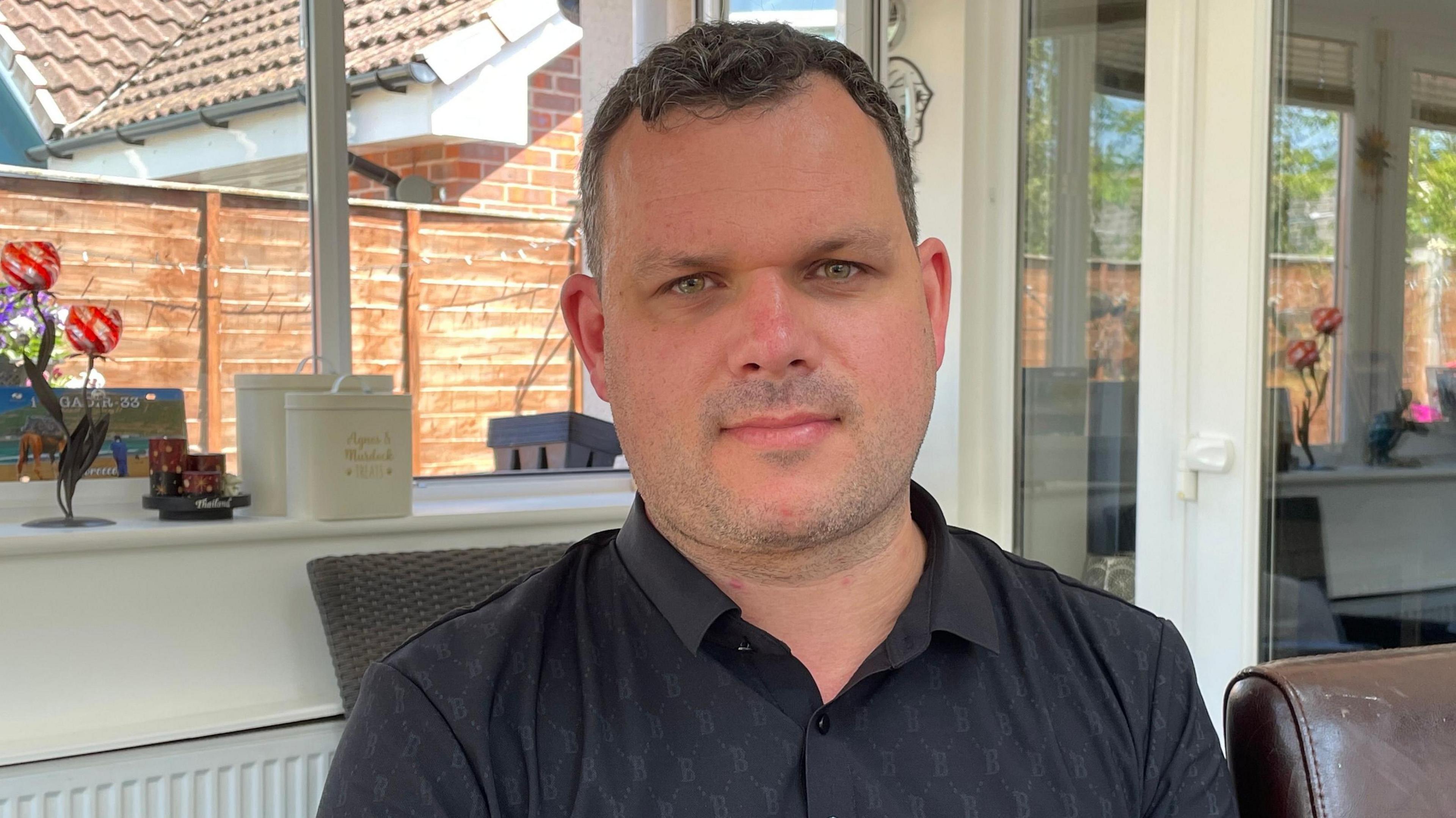 Ben Kingscote is pictured in front of a window. He is wearing a black button-up shirt and has short brown hair