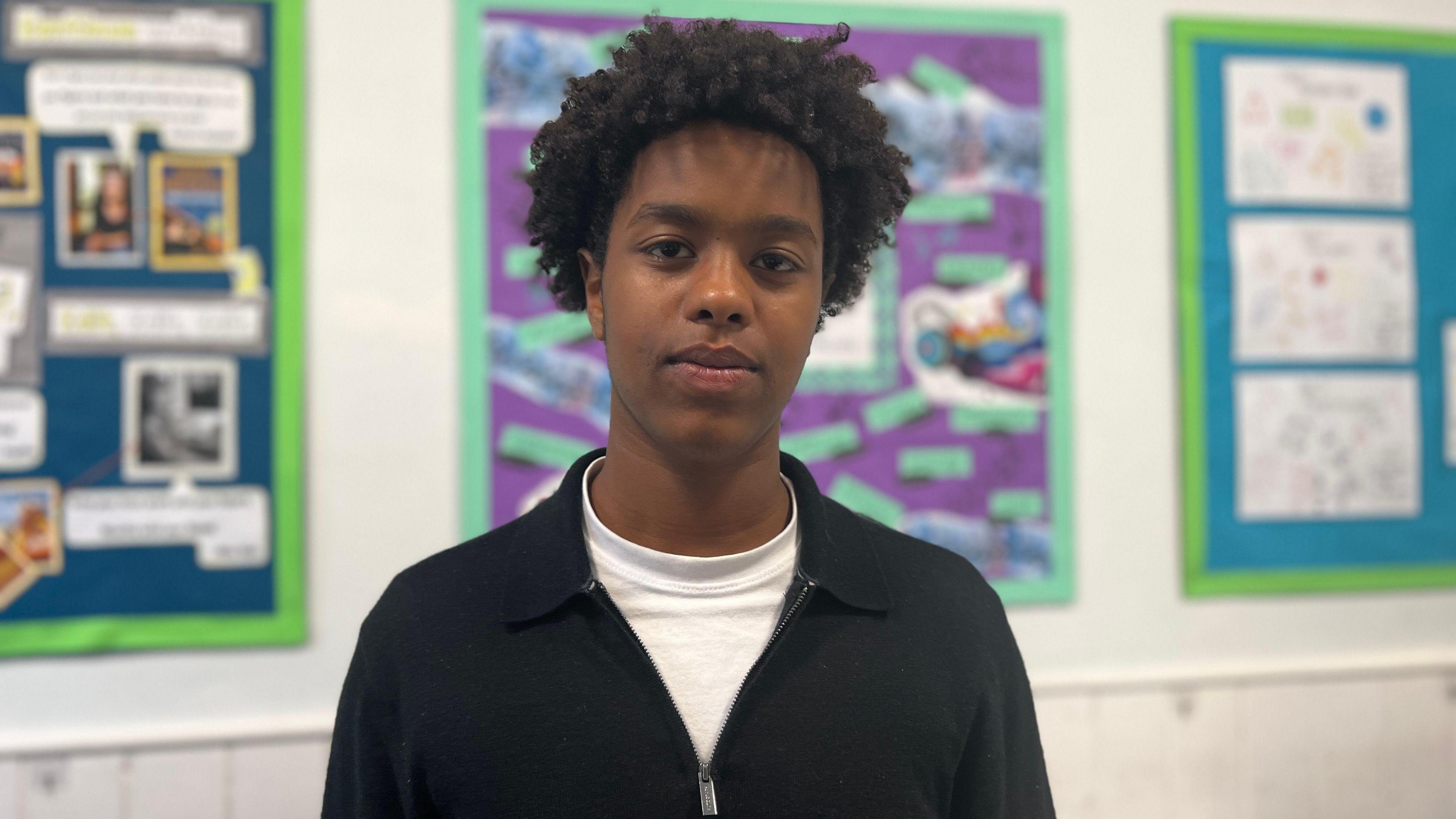 Ridwan. He has short, dark curly hair. He is wearing a white t-shirt and a black jumper. He is looking at the camera and smiling. 