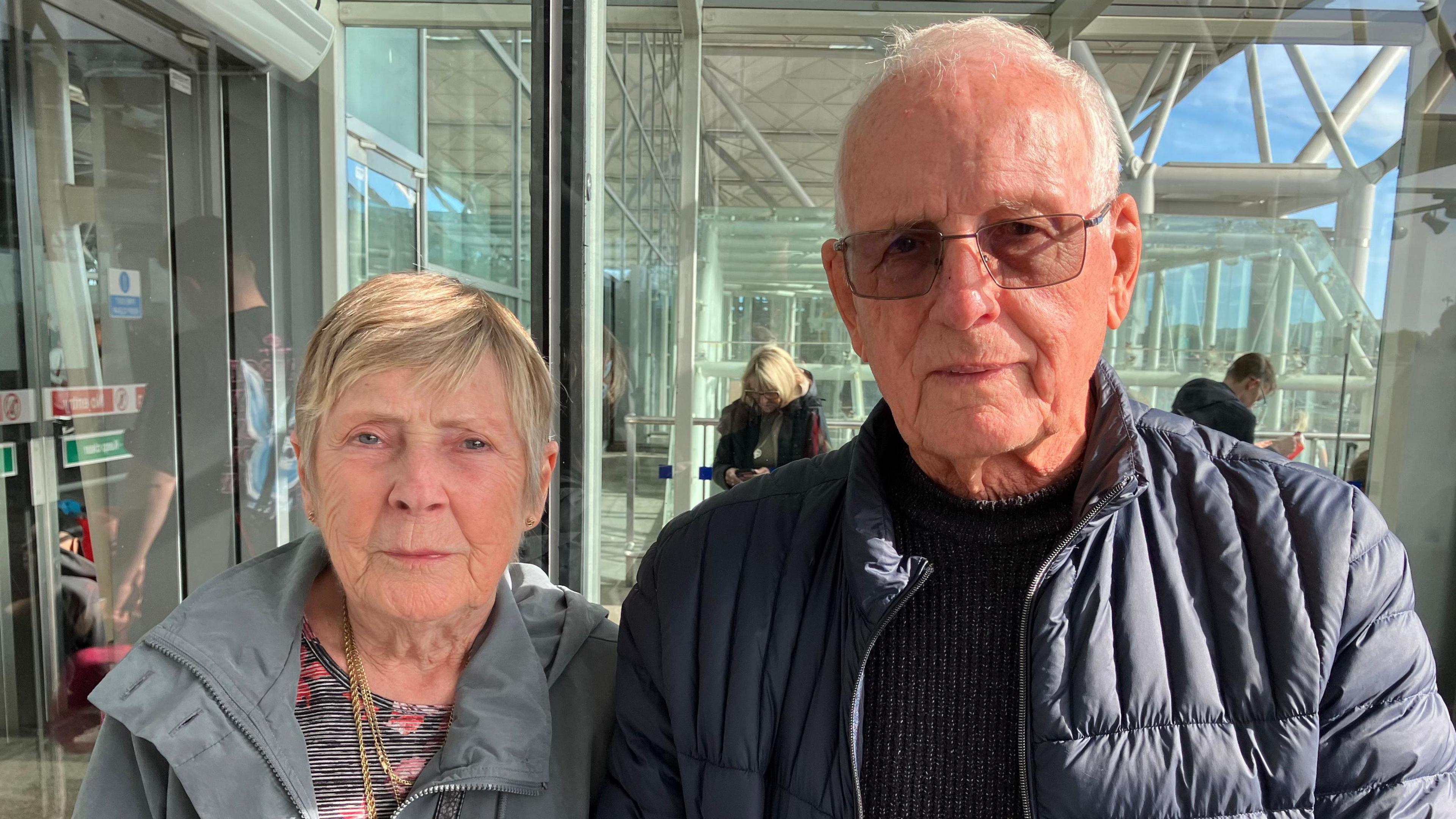 A lady and a man are stood next to each other. The lady, on the left, has short blonde hair and is wearing a floral top, a grey coat and a gold necklace. On the right is a man who has short grey hair, and is wearing a navy puffer jacket and black jumper.