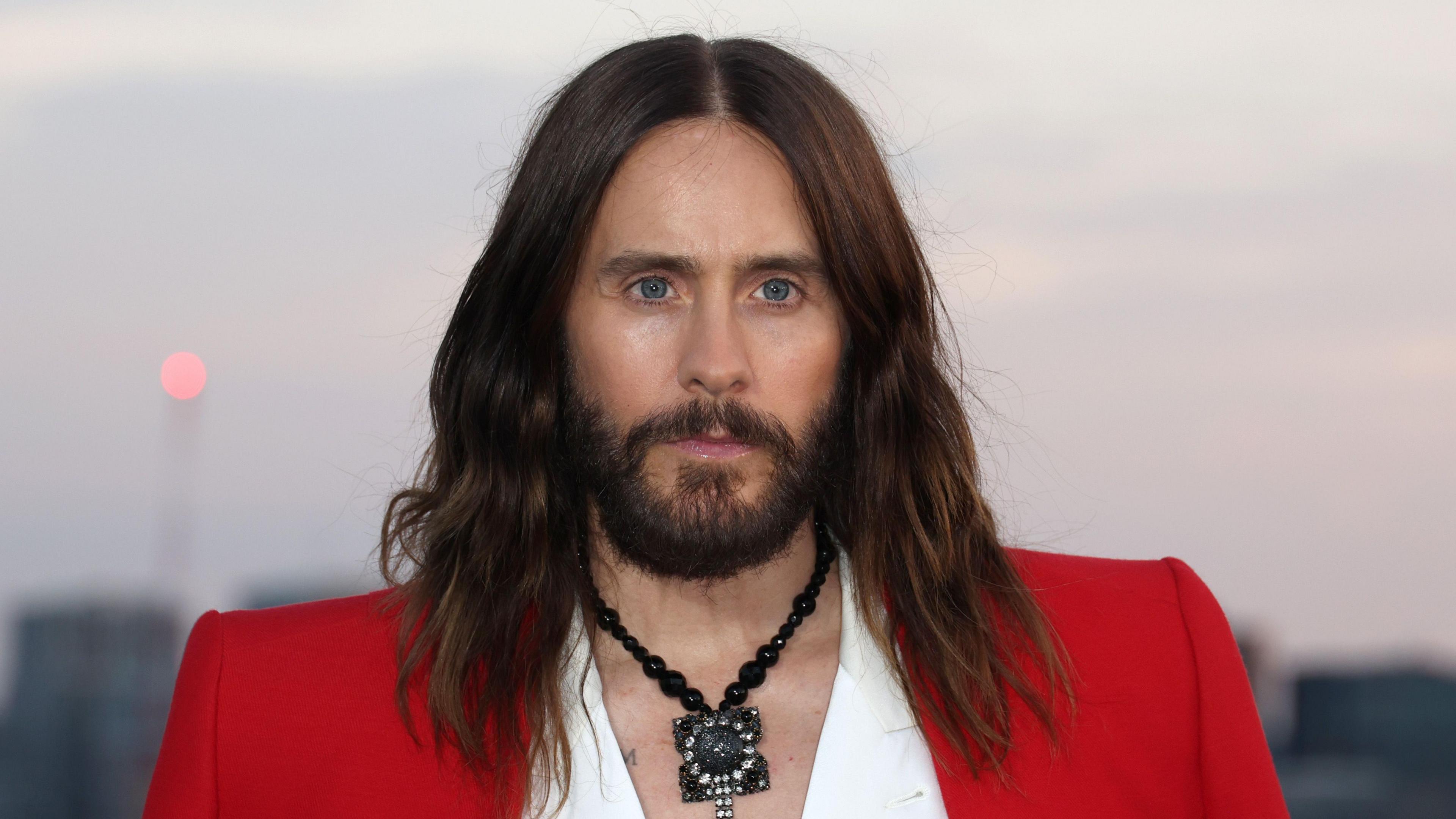 Jared Leto wears a red jacket over a white shirt. There is a large black necklace around his neck. 