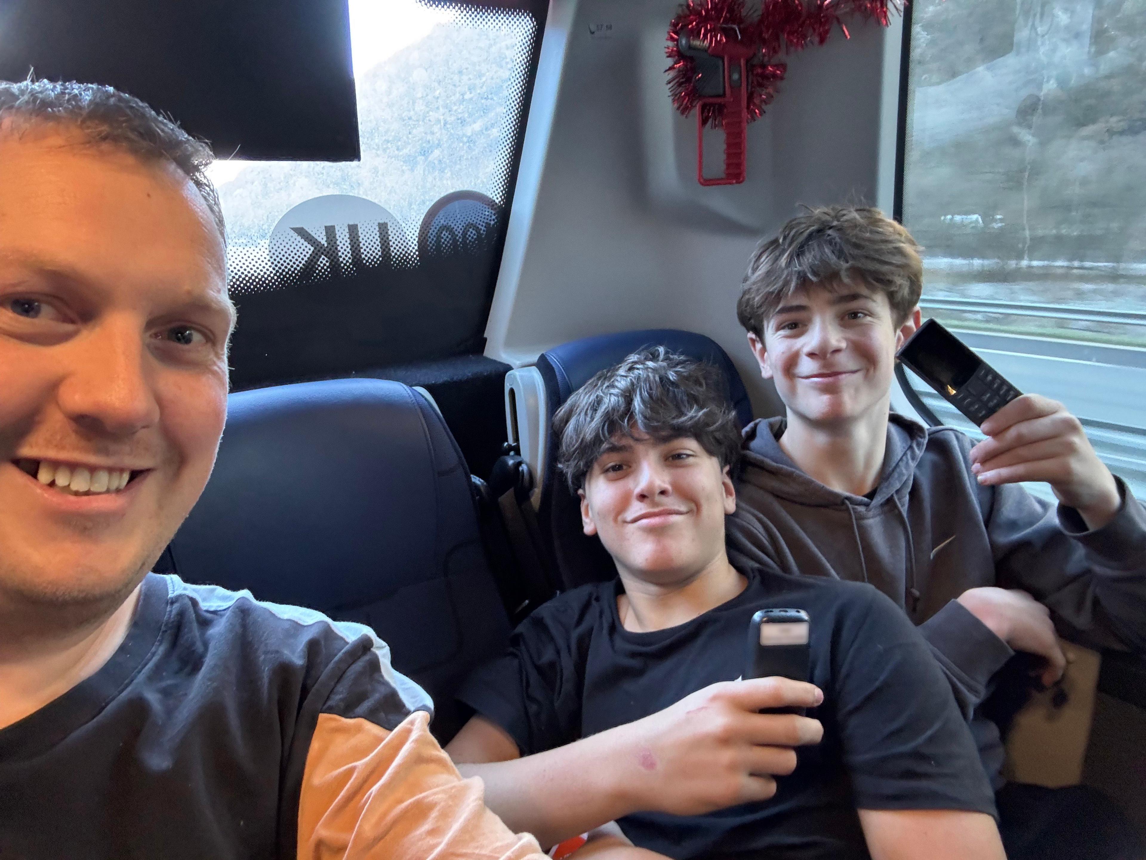 David Smith takes a selfie with two students, who are holding their brick phones and smiling, at the back of a school coach. 