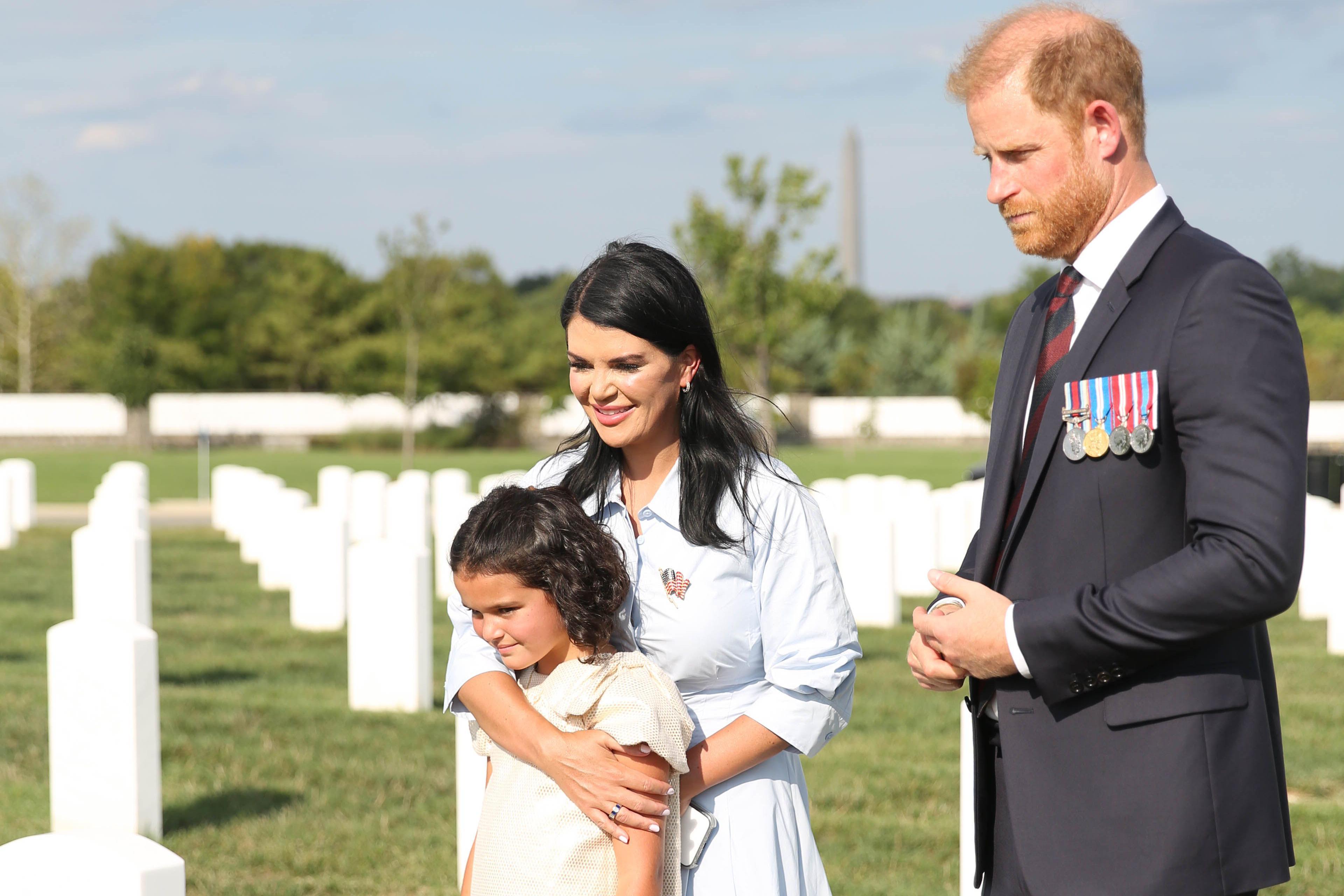Prince Harry stands in a suit with military medals next to a mother who has one arm around her young daughter. They are standing on a green graveyard honouring military personnel with white gravestones.