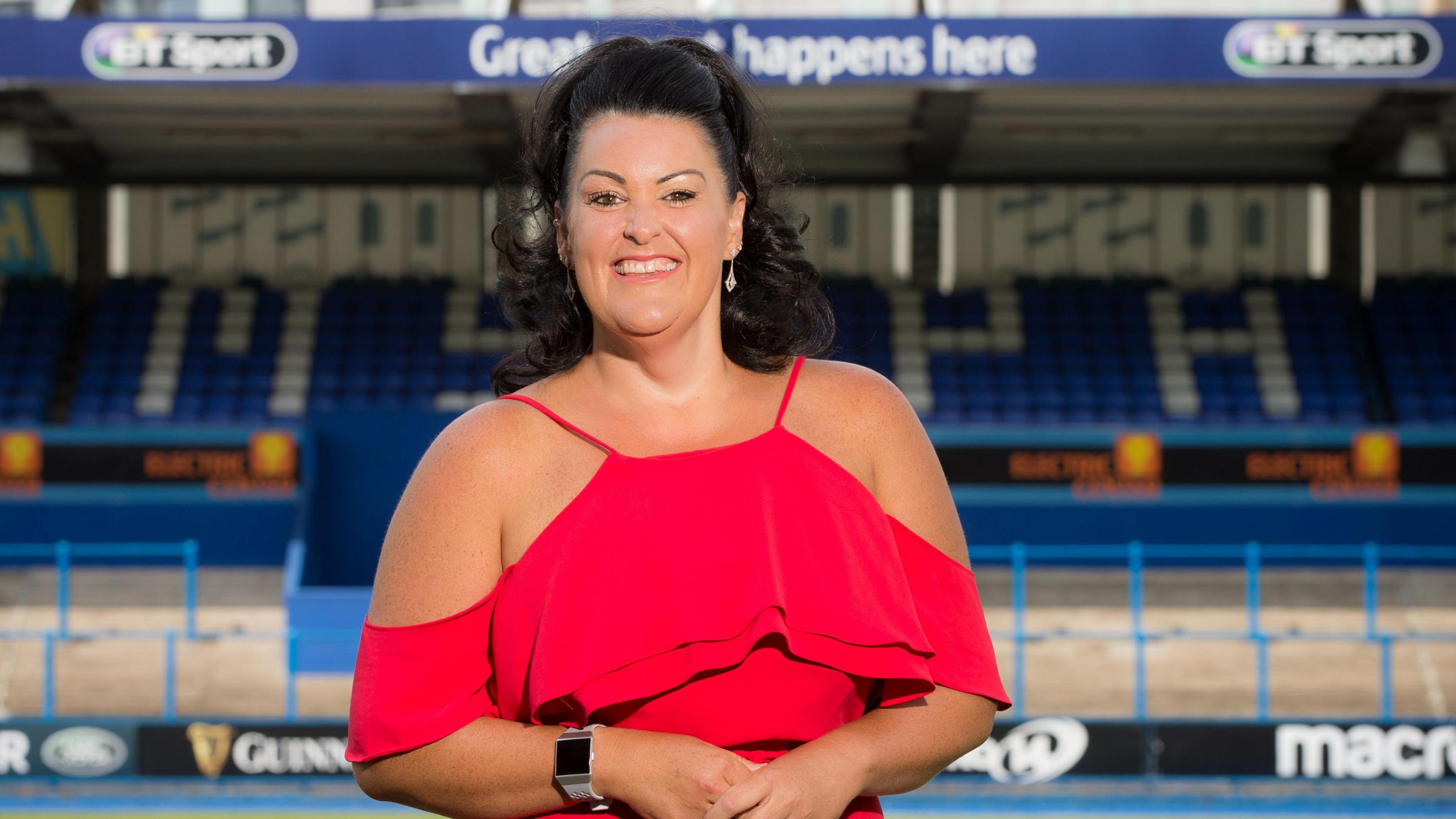 Hayley Parsons smiles at the camera in stood in the Cardiff Rugby stadium. She wears a red dress and drop earrings and her long dark hair is tied back. Blue seats can be seen in the stand behind her.