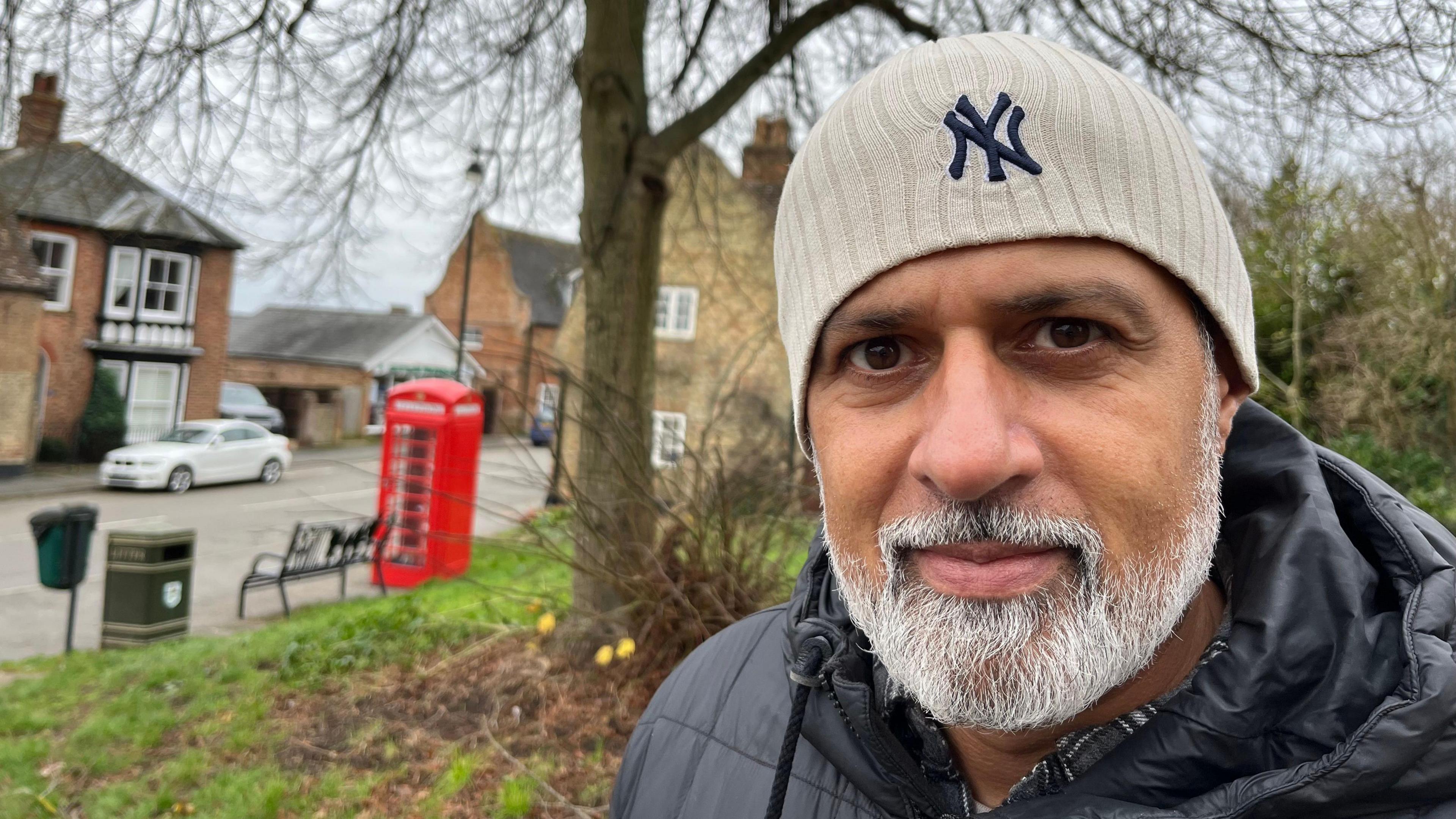 A close up of Mubeen as he is stood outside. He is wearing a black jacket and grey hat and has a white beard. Behind him there is a small patch of grass with a bench, bin and red telephone box. 