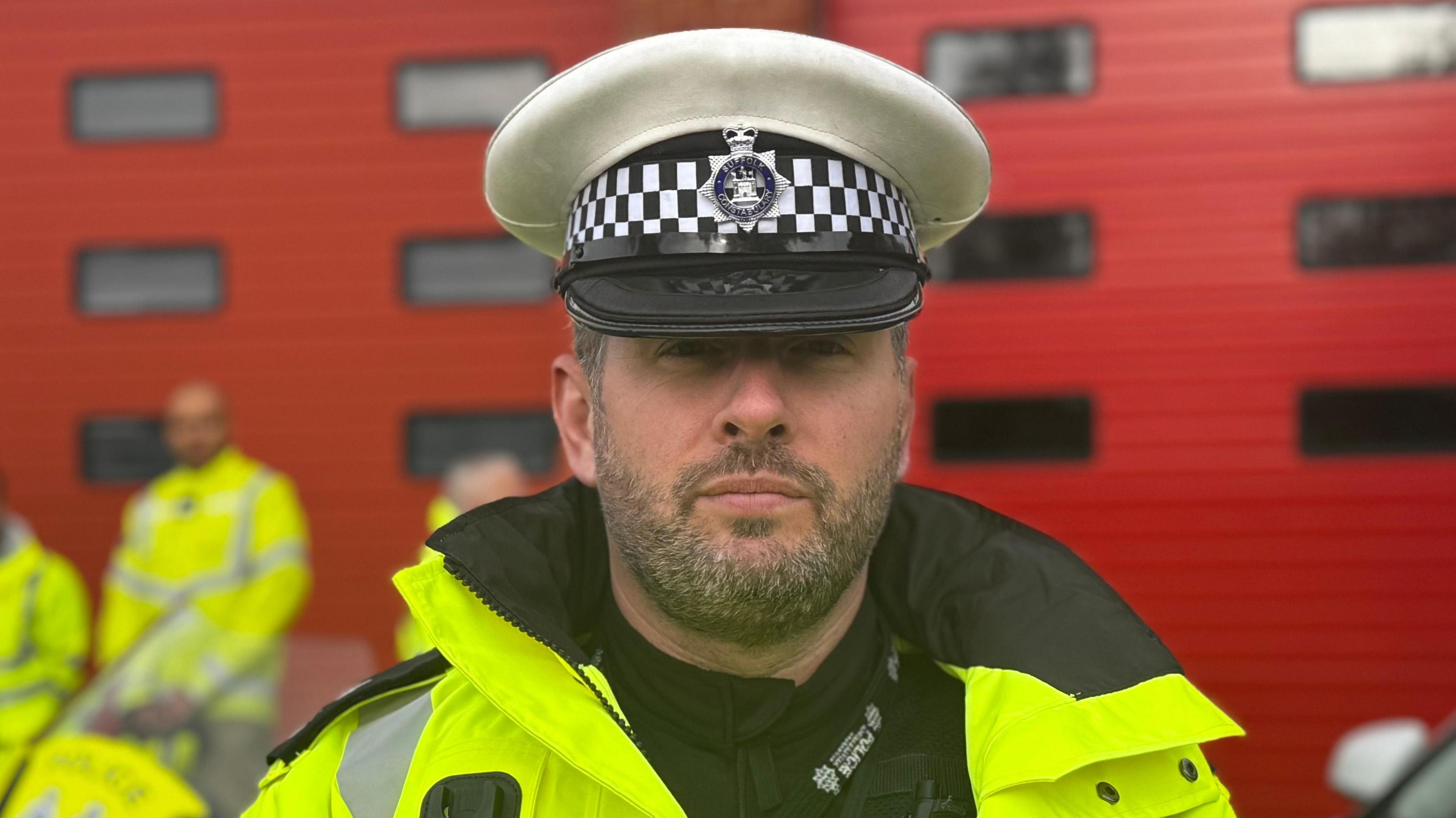 Bryon Hearsum looks directly at the camera with a solemn expression. He is wearing a high-vis jacket over a police uniform and a police hat. He is standing in front of two red doors.