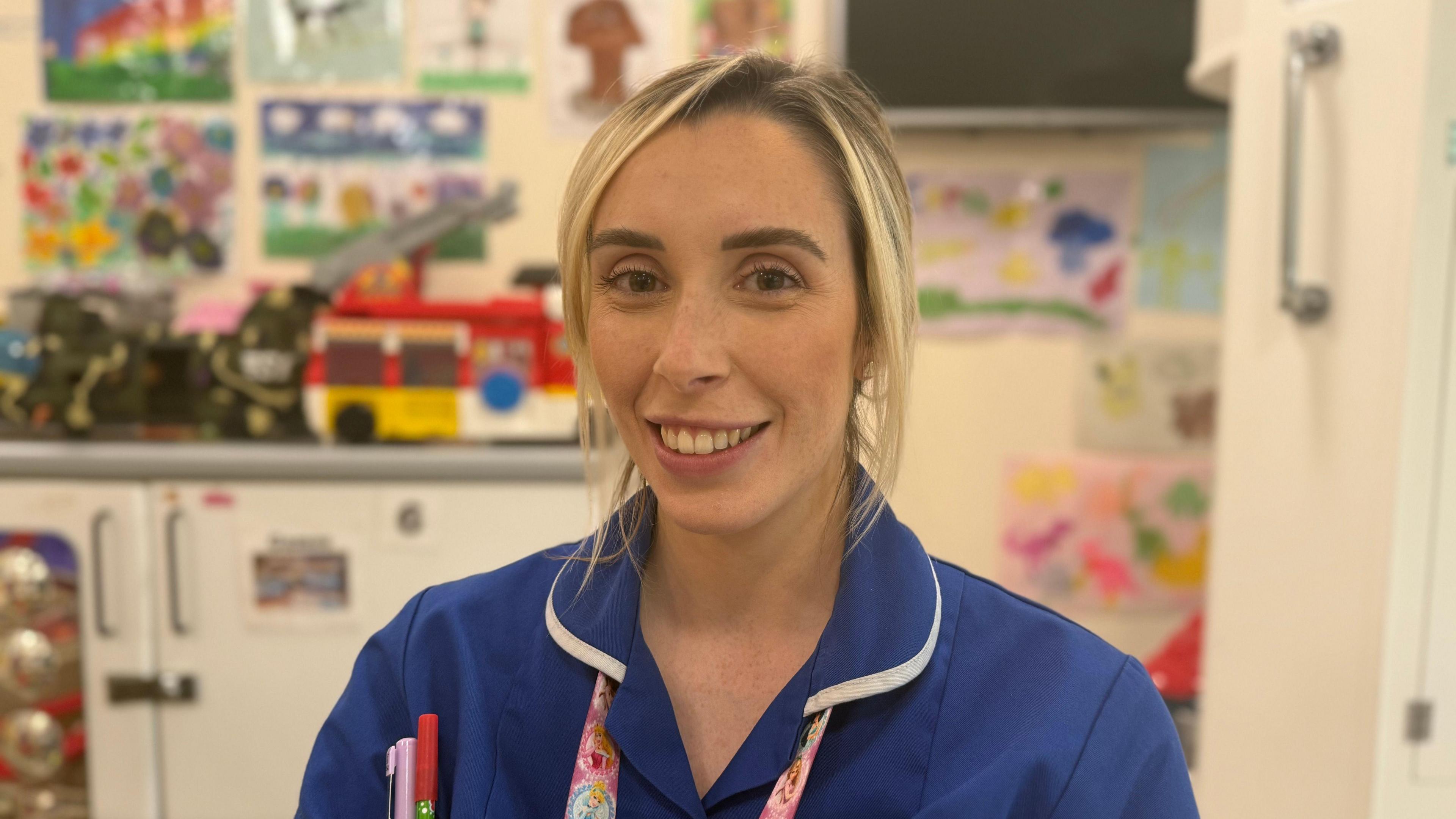 A woman with blonde hair, pulled back, smiles at the camera. She is wearing a blue nurse's uniform with a pink Disney princess lanyard. There are two pens in one of her pockets. She is standing in a playroom which has paintings on the walls. There are also children's toys in the background including a fire truck.