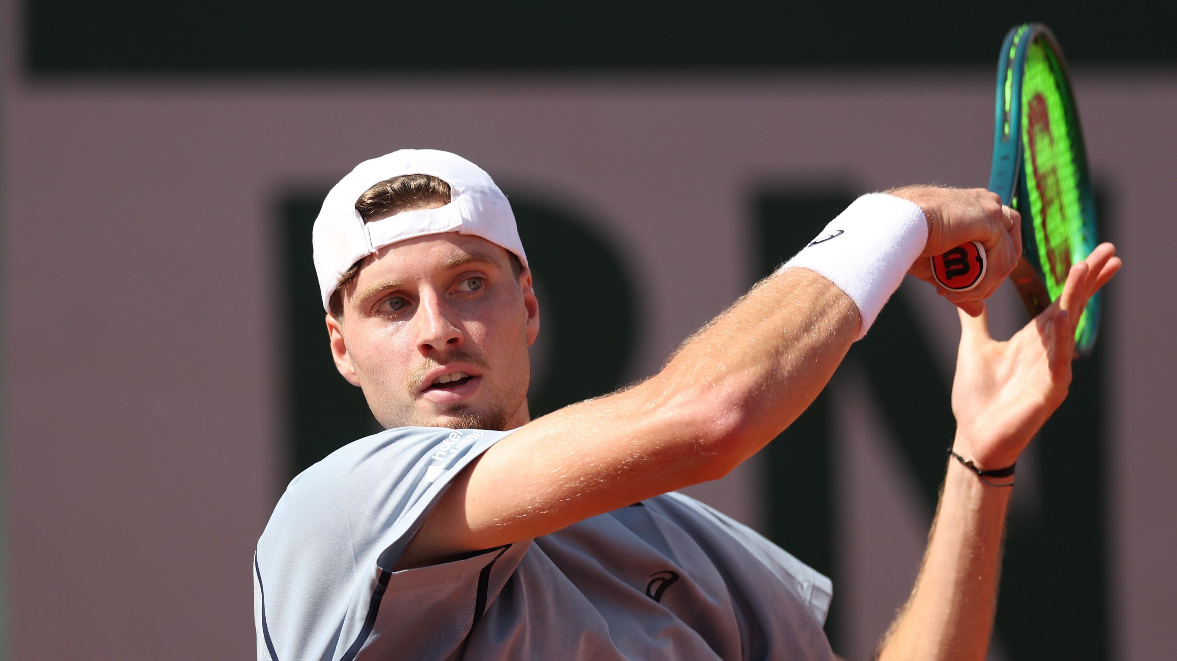 British tennis player Toby Samuel playing a forehand during a match at the French Open in Paris. Samuel is wearing a grey shirt and white hat, with the visor back to front. 