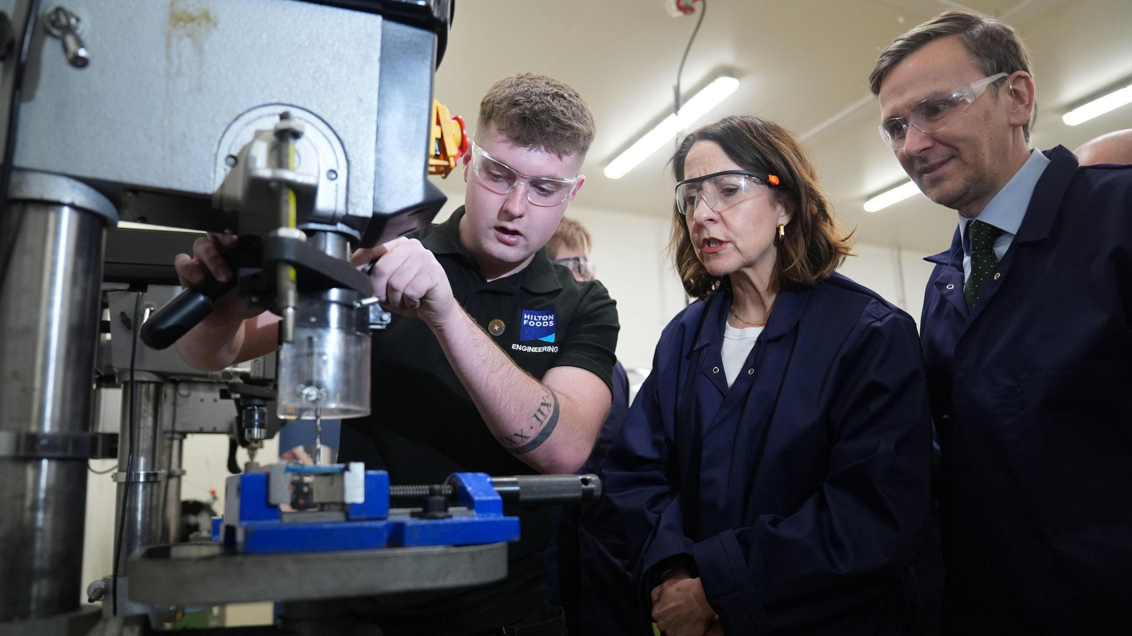 Work and Pensions Secretary Liz Kendall and Labour MP for Peterborough Andrew Pakes watch a young apprentice using a machine during a visit to Peterborough College. They are wearing safety goggles and overalls. 