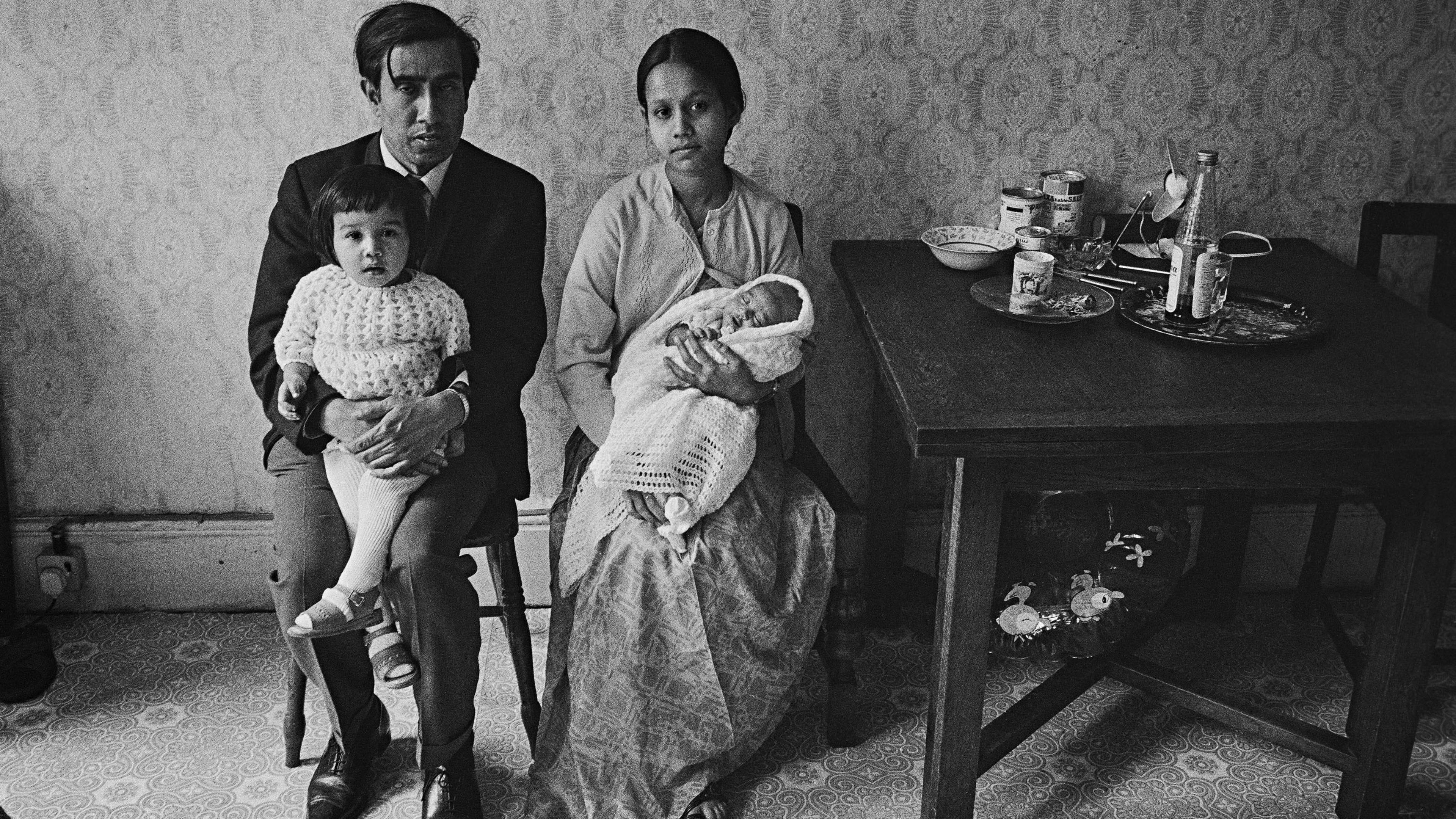Black and white image of a couple from Bangladesh facing the camera, sitting in a room by a table. The man is holding a small child in his arms. 