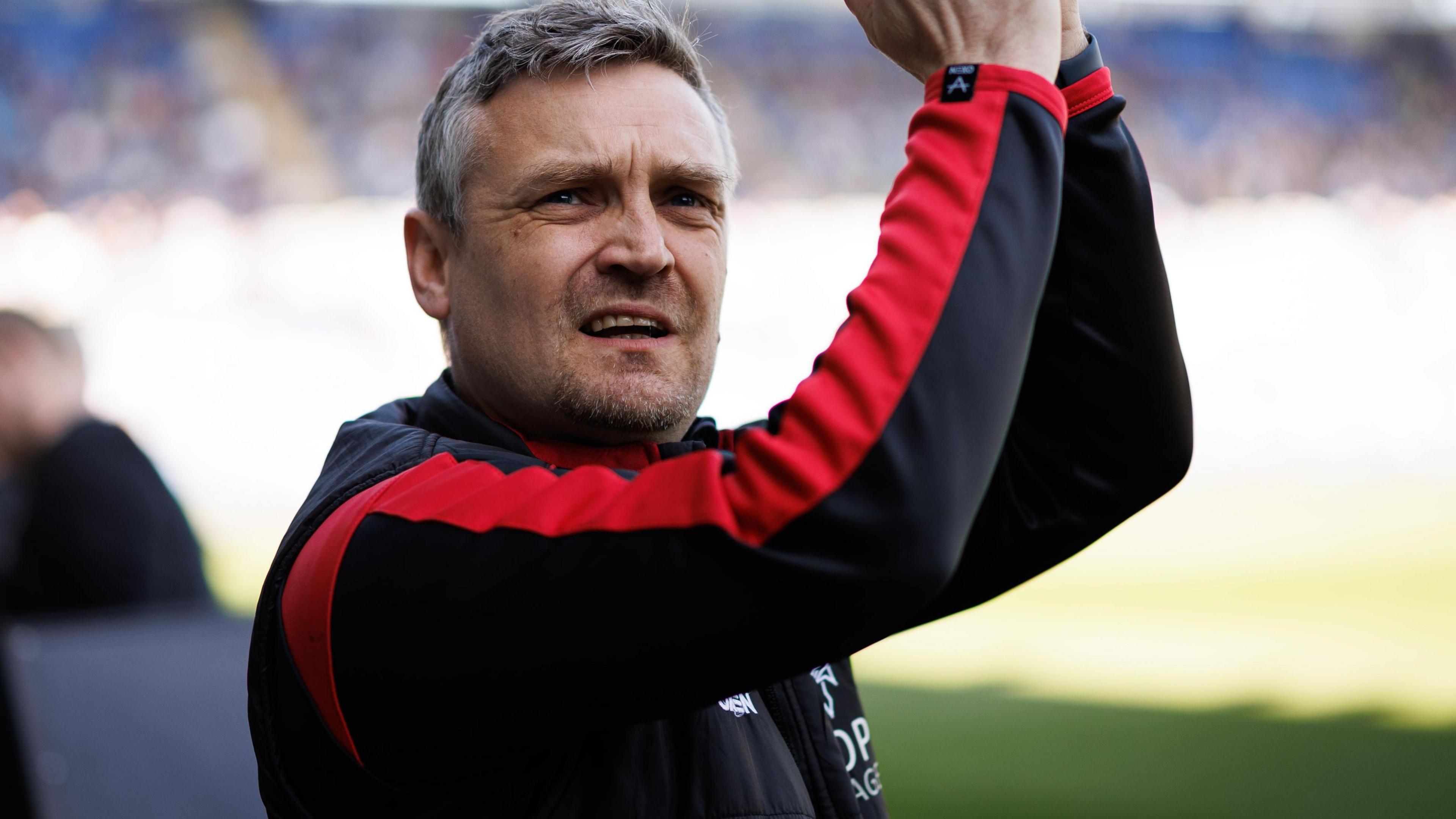 Bristol City manager Michael Skubala applauding fans while managing Lincoln City. Skubala is wearing a black training top with red sleeves.