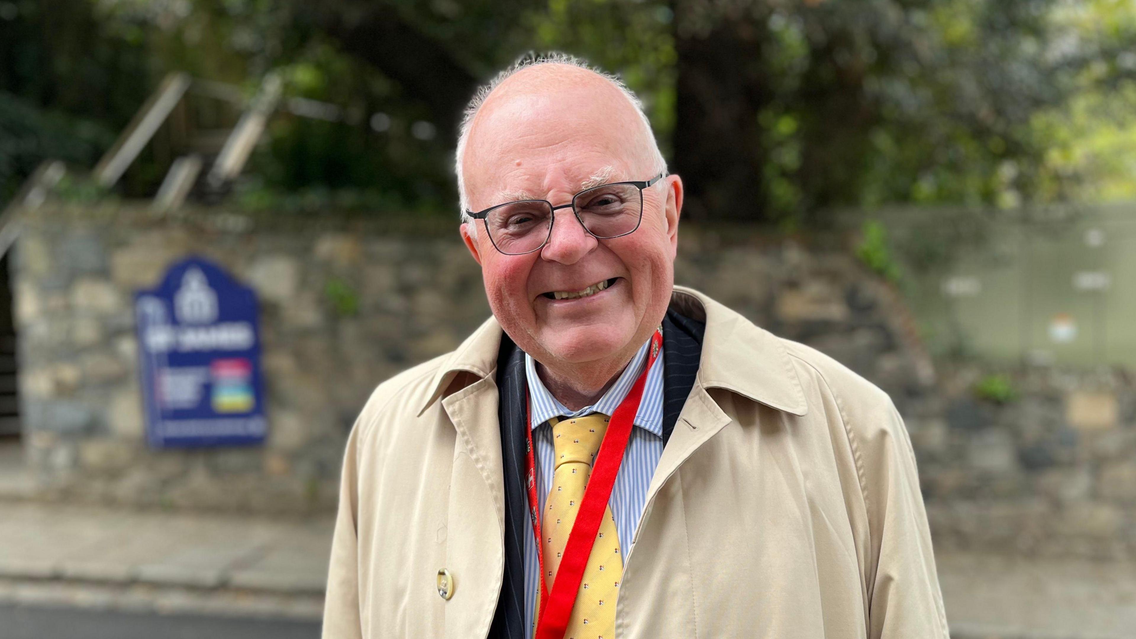 Deputy Chris Le Tissier - A bald man with very thin white hair on the side of his head. He's wearing a brown jacket, blue suit and a blue and white striped shirt, with a yellow tie that has blue dots on it. he has a red lanyard around his neck and is smiling towards the camera, wearing a black pair of glasses.